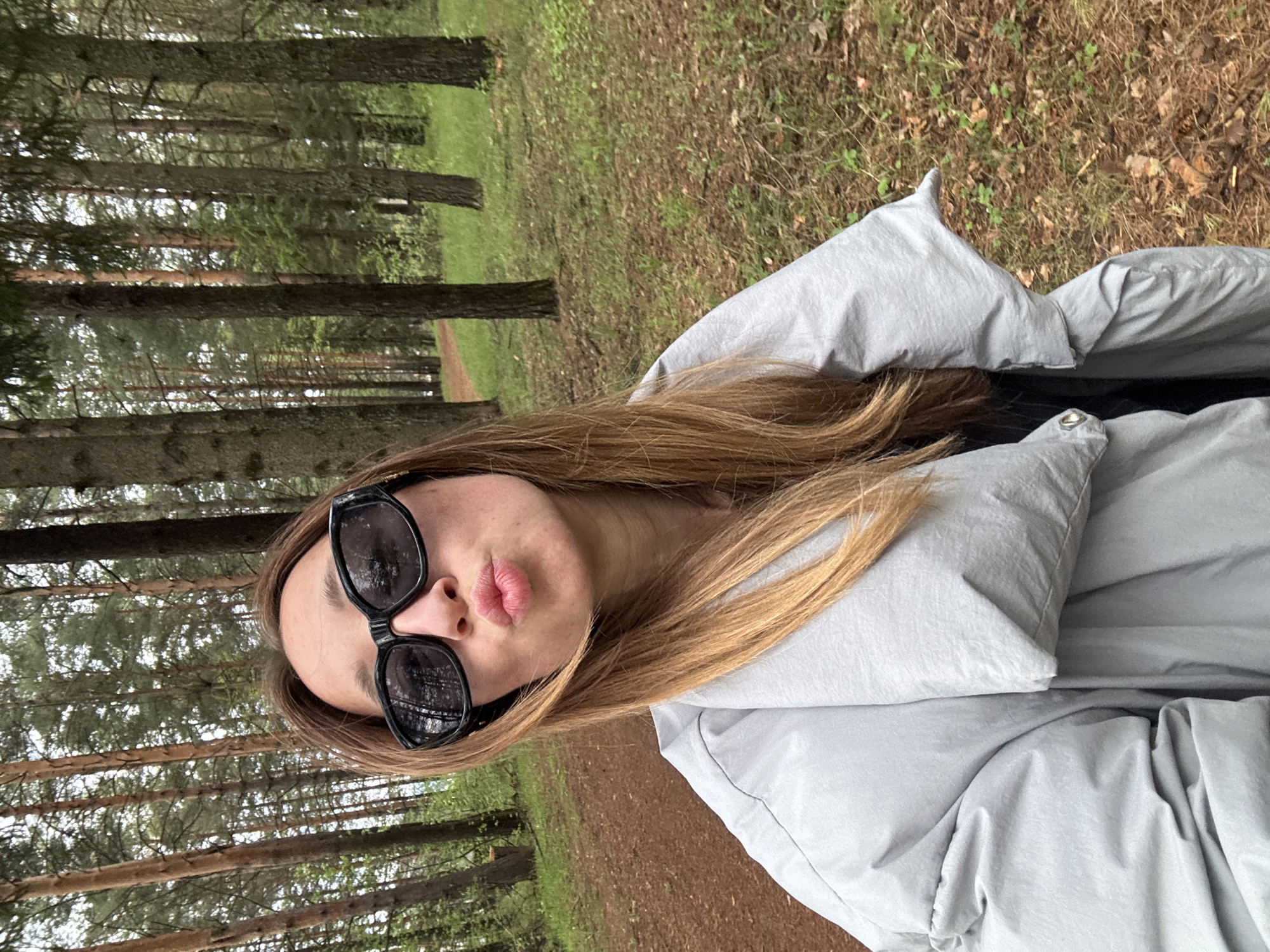 A young woman is posing for a selfie in a forest. She wears black sunglasses and a light gray puffer jacket. Her brown hair is long and partially covers the jacket. She's making a kissing face. The background shows trees and a forest floor covered in leaves and moss. The location is consistent with a wooded area in the city of Прикамский, Russia.