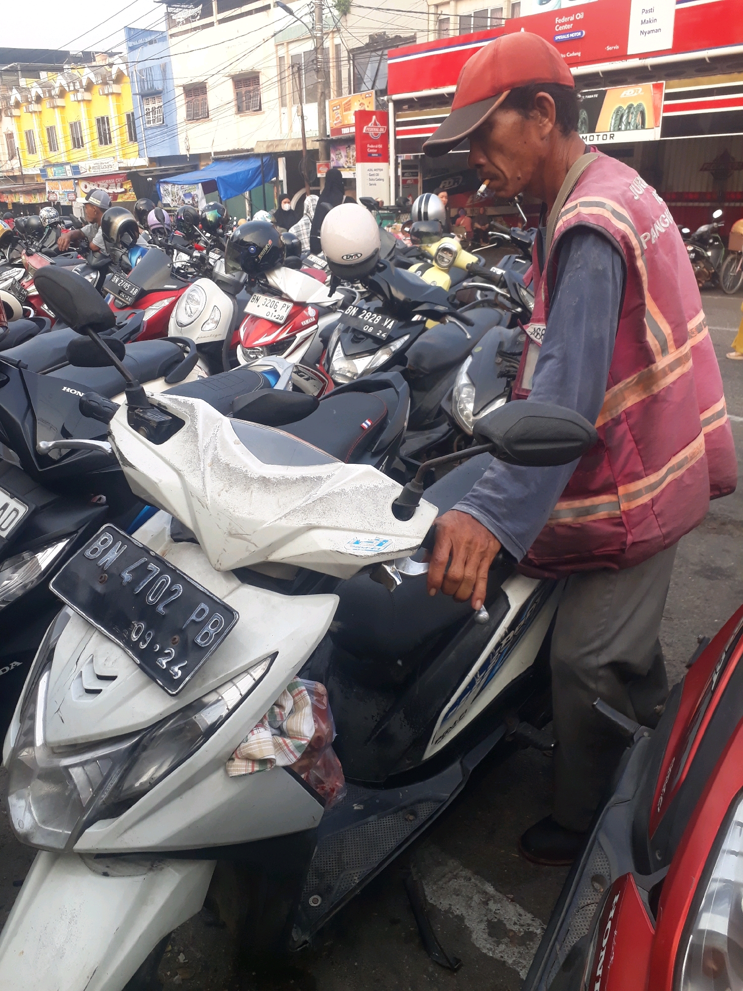 A middle-aged man wearing a red cap and a red reflective vest over a blue long-sleeved shirt, is smoking a cigarette while standing next to a white scooter. The scooter has a license plate with the inscription "BN 4702 PB" and "09.24". The scene appears to be a street in Pangkalpinang, Indonesia, with a row of parked motorcycles and scooters in the background. The buildings in the background suggest an urban environment. The man's posture and the presence of a cigarette indicate a momentary pause or break. The overall impression is a snapshot of daily life in a busy Indonesian city.