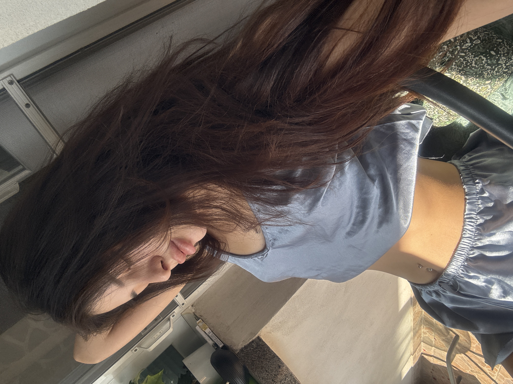 A sunlit, portrait-style shot, tilted horizontally, features a young woman with long, dark brown hair that flows down around her. Her head is tilted, showing her face in profile with her lips slightly parted and a relaxed expression. She is wearing a light blue or grey silky tank top and matching shorts, resembling pajamas or loungewear. Two small silver studs of a belly button piercing are visible on her midriff.
The scene is brightly illuminated by natural sunlight, indicating it is daytime, likely morning or late afternoon, as strong shadows are cast across the surfaces. The setting appears to be an indoor space near a window or on a balcony. To the left, a window with a grey mesh screen and a metal frame is visible. On the right side, behind the woman, parts of a dark chair and green patterned fabric are present, along with glimpses of green foliage from plants. A light-colored, possibly beige, exterior wall of a building is also part of the background. The overall impression is one of ease and comfort in a residential or similar outdoor-facing setting in Plovdiv, Bulgaria. No visible text is present in the image.