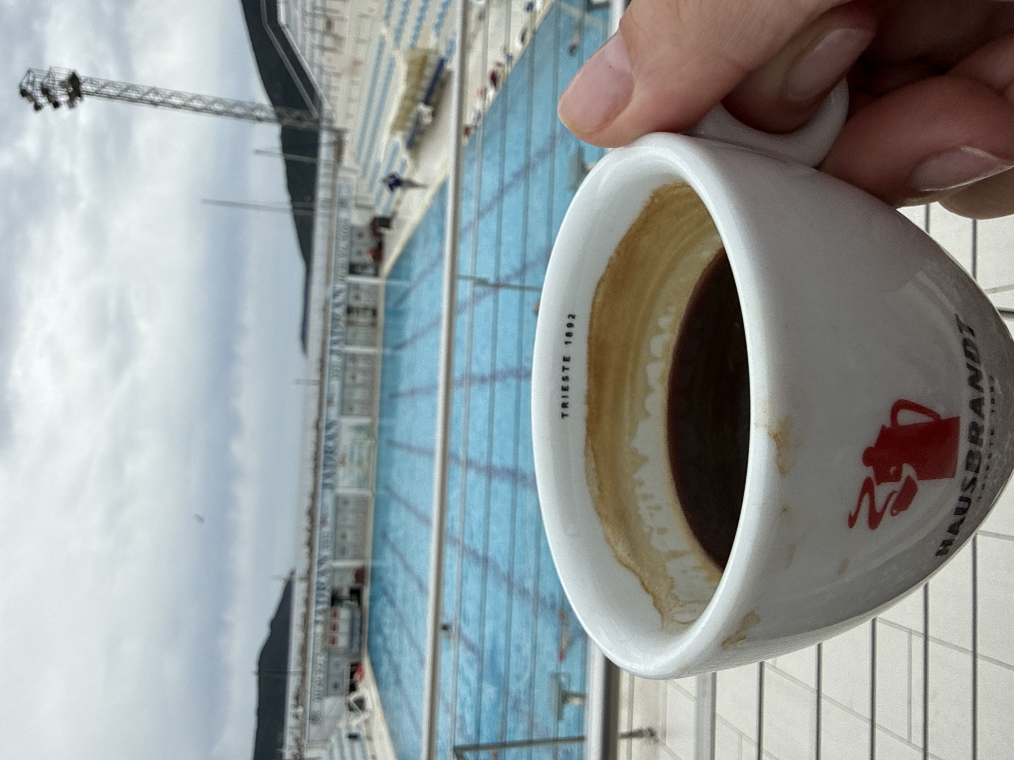 A tilted, medium shot captures a partially consumed cup of coffee held in a person's hand with a background of an outdoor swimming pool area. The cup is white with a red logo featuring a coffee pot and the word "HAUSBRANDT." "TRIESTE 1892" is printed inside the lip of the cup. The pool area, with blue lanes and rows of empty seats, is located in Herceg Novi, Montenegro. A tall metal lighting tower is visible in the background. The sky is overcast. The foreground shows a light-colored tiled surface, likely a balcony or terrace.