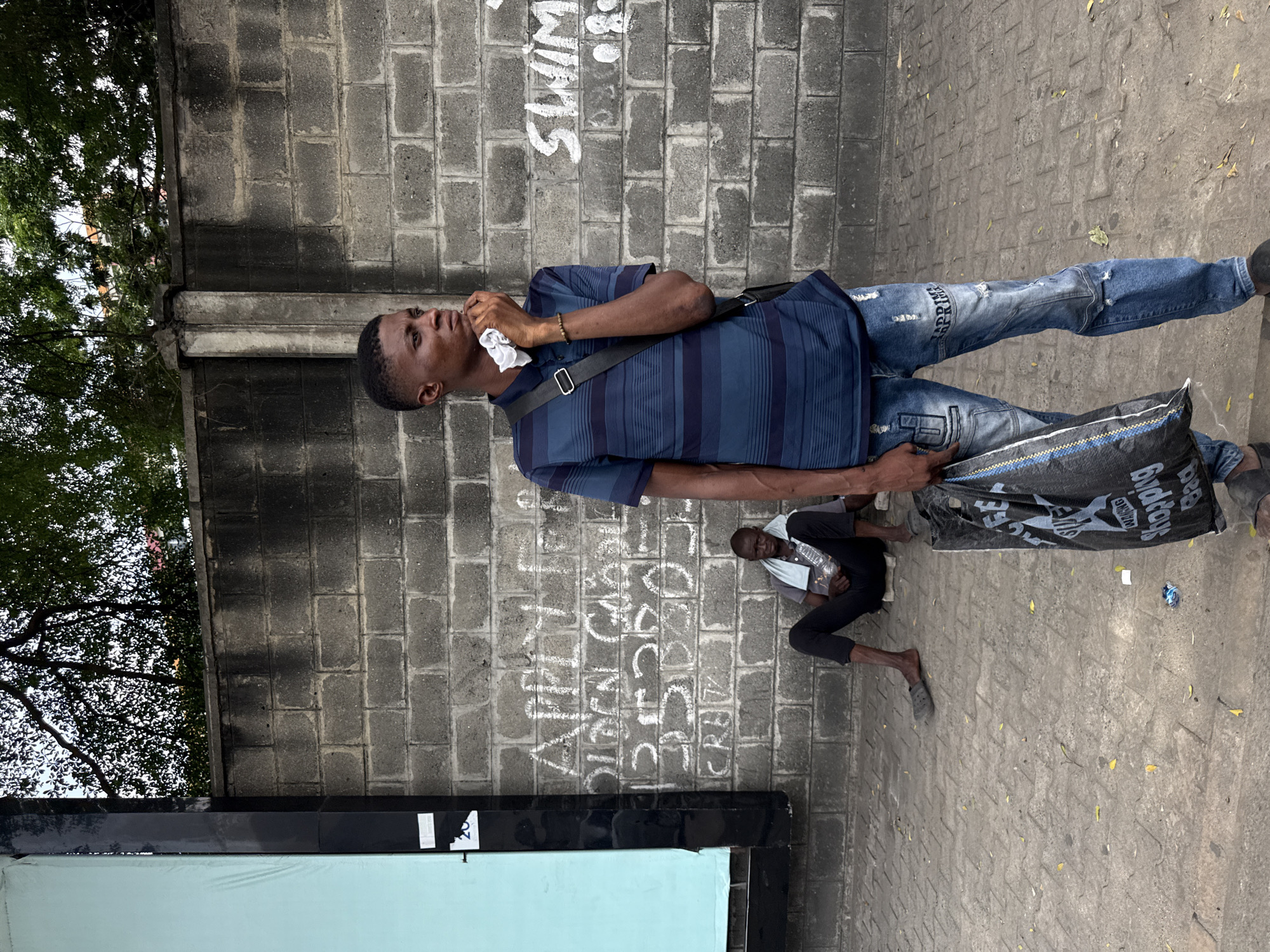 A man is standing on a sidewalk in Itirin, Nigeria. He is wearing a blue striped shirt, jeans, and a black strap across his chest. He is holding a crumpled white cloth to his face and has a black bag in his other hand. Behind him is a cinderblock wall with graffiti and another man sitting on the ground, wearing black pants and a light-colored shirt. The sidewalk is paved with gray bricks and there are trees visible in the background.
