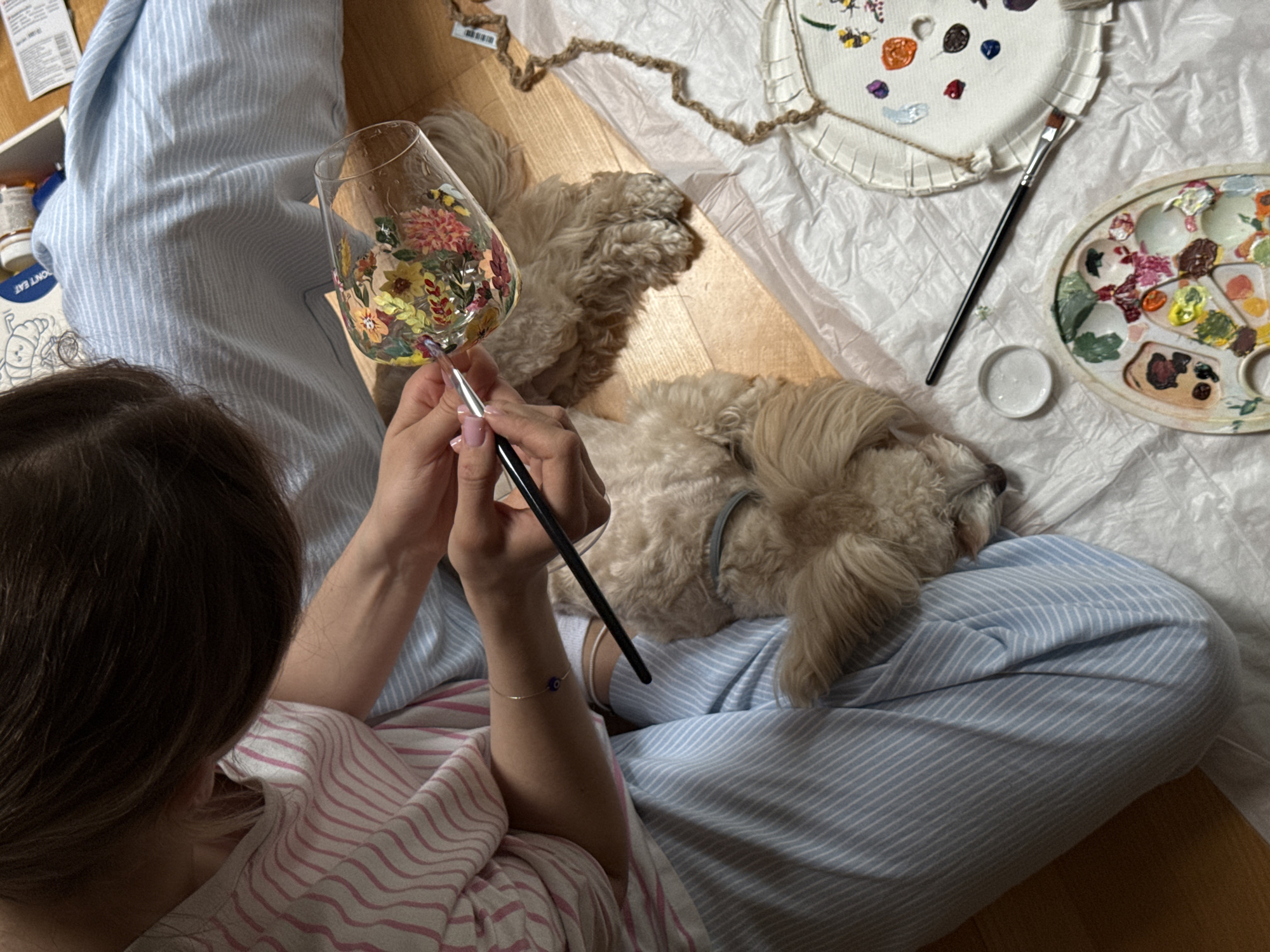 A person with brown hair and a pink and white striped shirt is sitting cross-legged, painting a wine glass with a floral design. They are holding the glass in their left hand and a paintbrush in their right. A light-brown dog is lying on their lap. Beside the person is a white plastic sheet with a palette of paints, a paintbrush, and a white disc-shaped canvas. The scene is indoors on a wooden floor. There is no direct evidence of the city Sapronovo, Russia.
