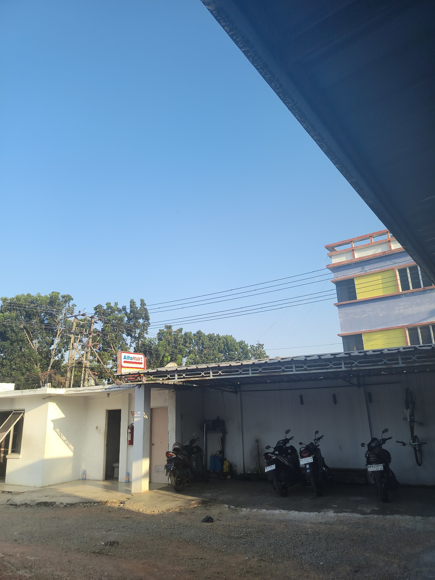 The image shows a bright, clear blue sky above a scene in Pangkalpinang, Indonesia. In the foreground, a dark, angled structure casts a shadow. To the left, a white building with a window and an open doorway is visible. Inside the doorway, a toilet is partially visible, and a red fire extinguisher is mounted on the wall. Orange and blue bottles are placed on the floor near the entrance. A sign for "Alfamart" is mounted on a pole above a covered walkway. This walkway extends to the right, sheltering several parked motorcycles. The motorcycles are dark in color, and their license plates are visible. To the right of the motorcycles, a bicycle is mounted on the wall. Behind the covered walkway, a multi-story building with yellow and blue sections is visible against the sky. Electrical poles and wires are also present in the scene. The ground is covered with gravel and dirt.