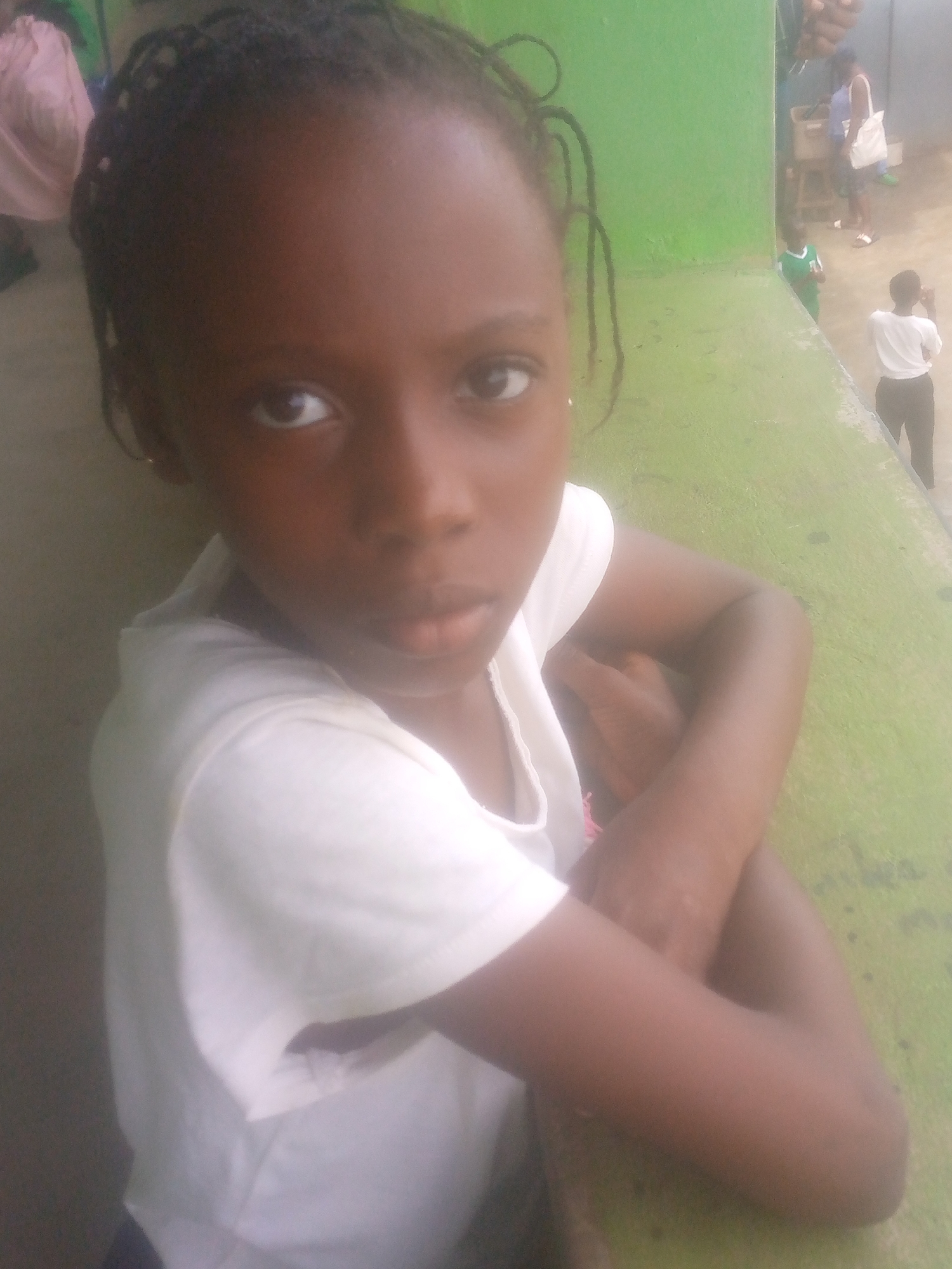 A young girl with braided hair, wearing a white t-shirt, is sitting on or leaning against a green surface. She has her arms crossed and is looking directly at the camera. In the background, there are other people, some wearing white shirts and dark pants. The scene appears to be outdoors, possibly near a building with a green exterior. The exact location within the city of Unknown, Unknown is not discernible from the image.