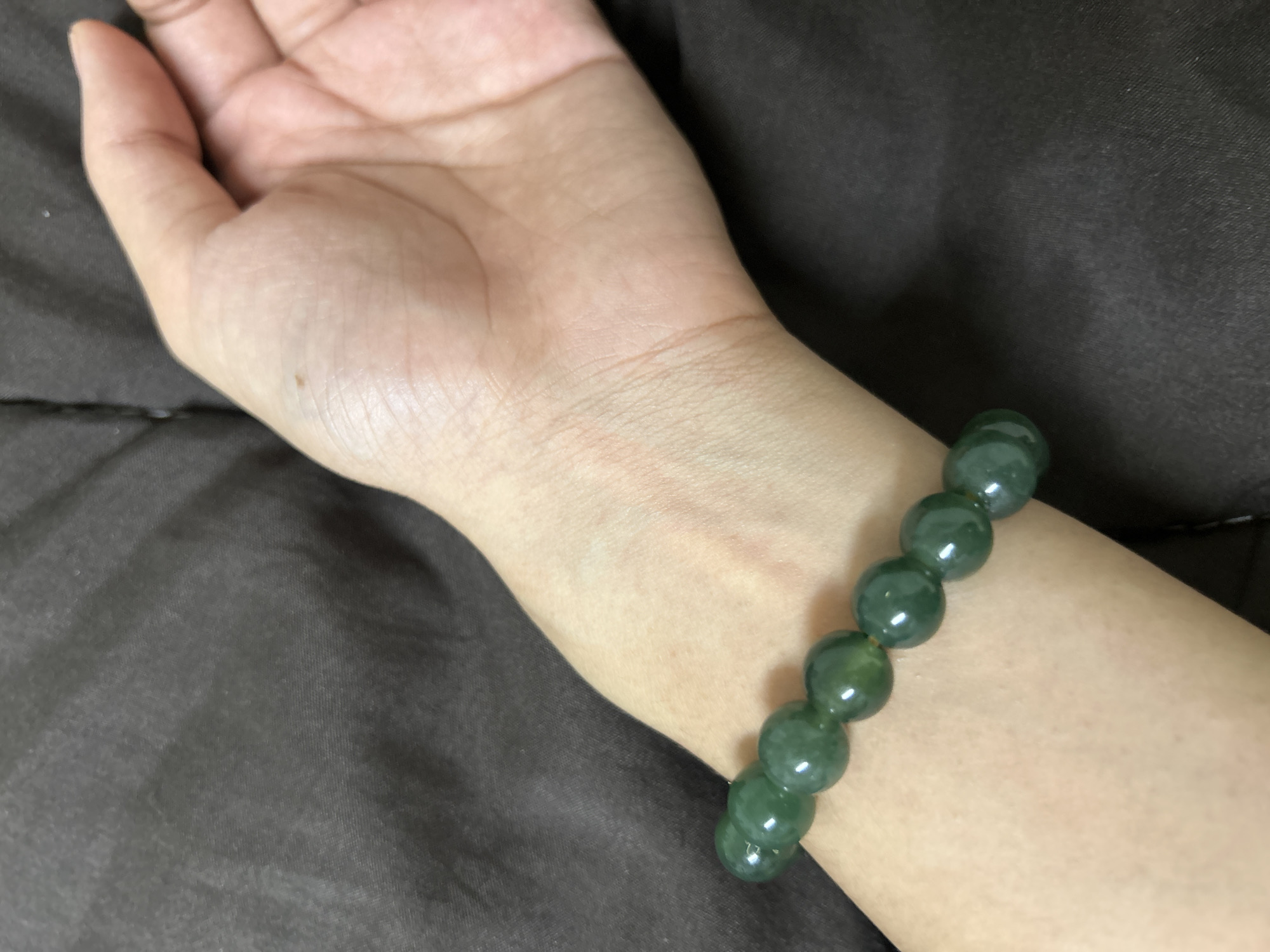 A close-up shot shows a human wrist adorned with a bracelet made of smooth, green, round beads. The wrist is resting on a textured, dark brown surface, possibly fabric. The lighting casts subtle shadows, highlighting the curvature of the wrist and the sheen of the beads. The background is out of focus, emphasizing the bracelet and wrist as the primary subjects. No specific location in Khlong Luang Town Municipality, Thailand, can be determined from this image.