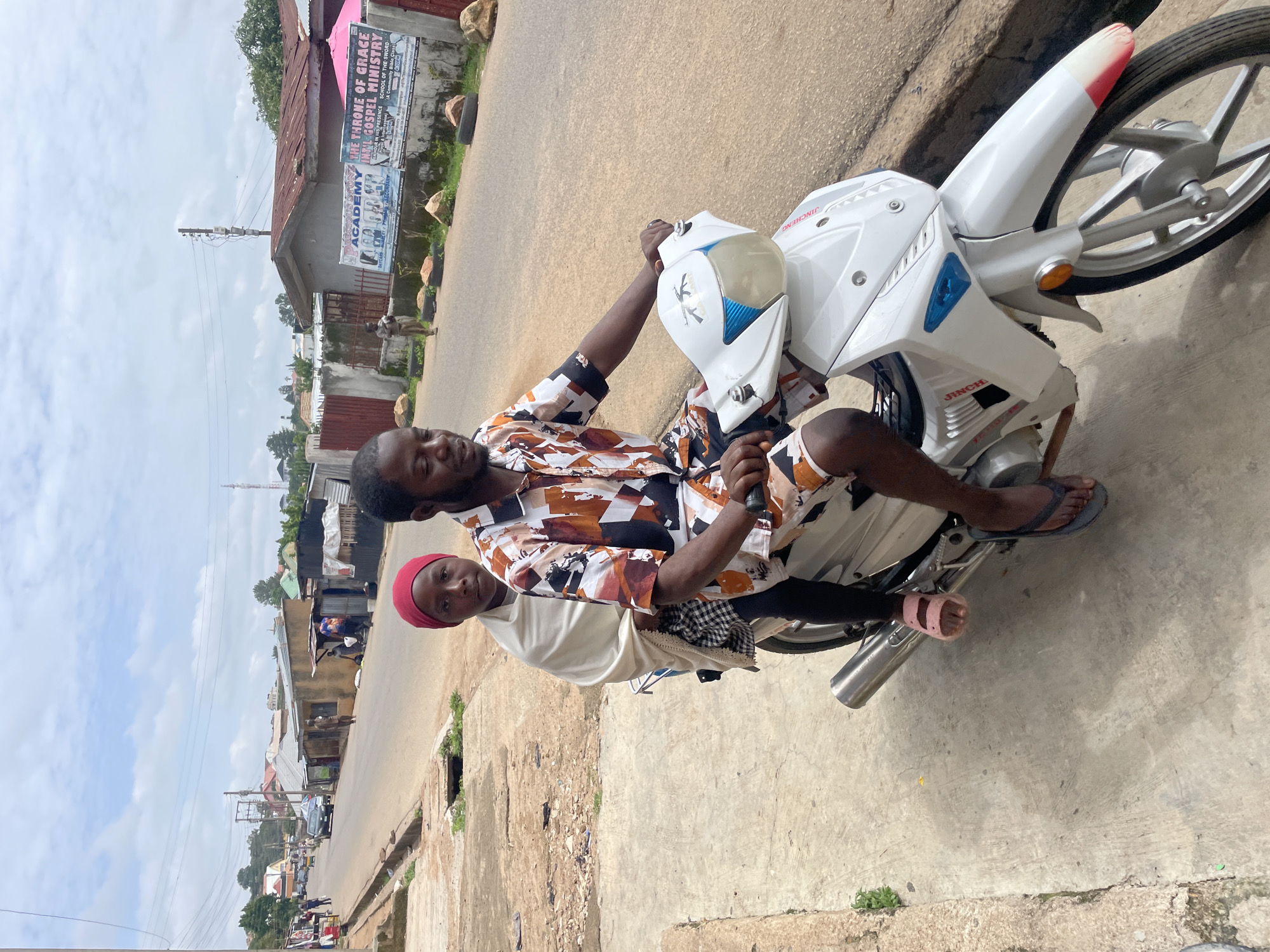 In Jos, Nigeria, on a paved road with a concrete sidewalk, a man and a young girl ride on a white motorcycle. The man, wearing a patterned short-sleeved shirt and shorts, drives the motorcycle. The girl, seated behind him, is wrapped in a white shawl and wears a red headscarf. The motorcycle has a silver exhaust pipe and a red and white accent on the front fender. In the background, buildings with corrugated metal roofs line the street, and a billboard for "Precious Stonez Academy" and "The Throne of Grace Intl Gospel Ministry" is visible on a wall. Several people walk along the road and sidewalk. The sky is partly cloudy.