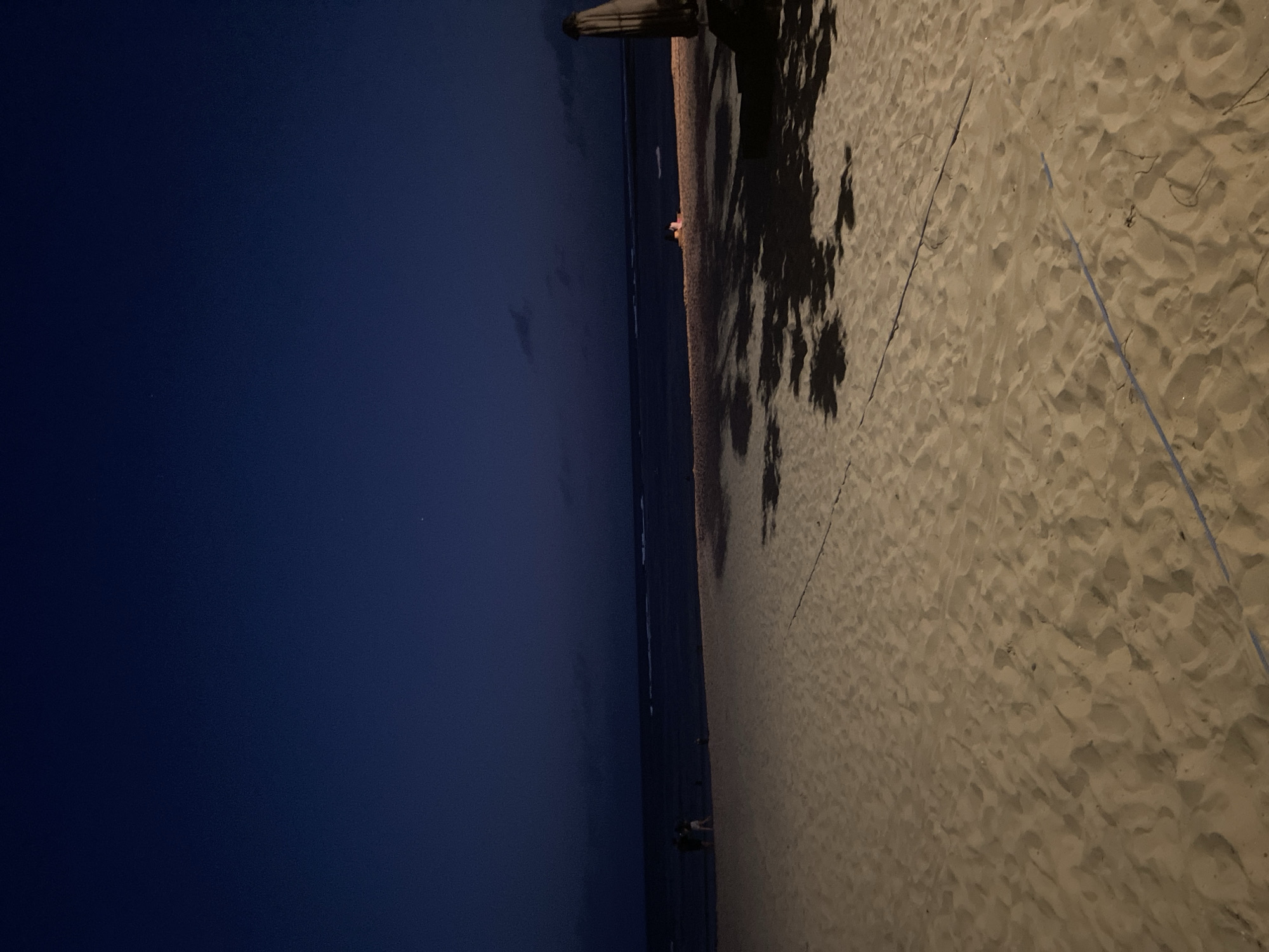The image depicts a beach in Kuta Selatan, Indonesia at night. The left side of the image shows a dark blue sky and ocean. The right side is a sandy beach. There are dark shadows on the sand, possibly from foliage. A blue line is visible running across the sand. In the distance, a small group of people are visible on the beach. There is a faint glow of light further along the beach.