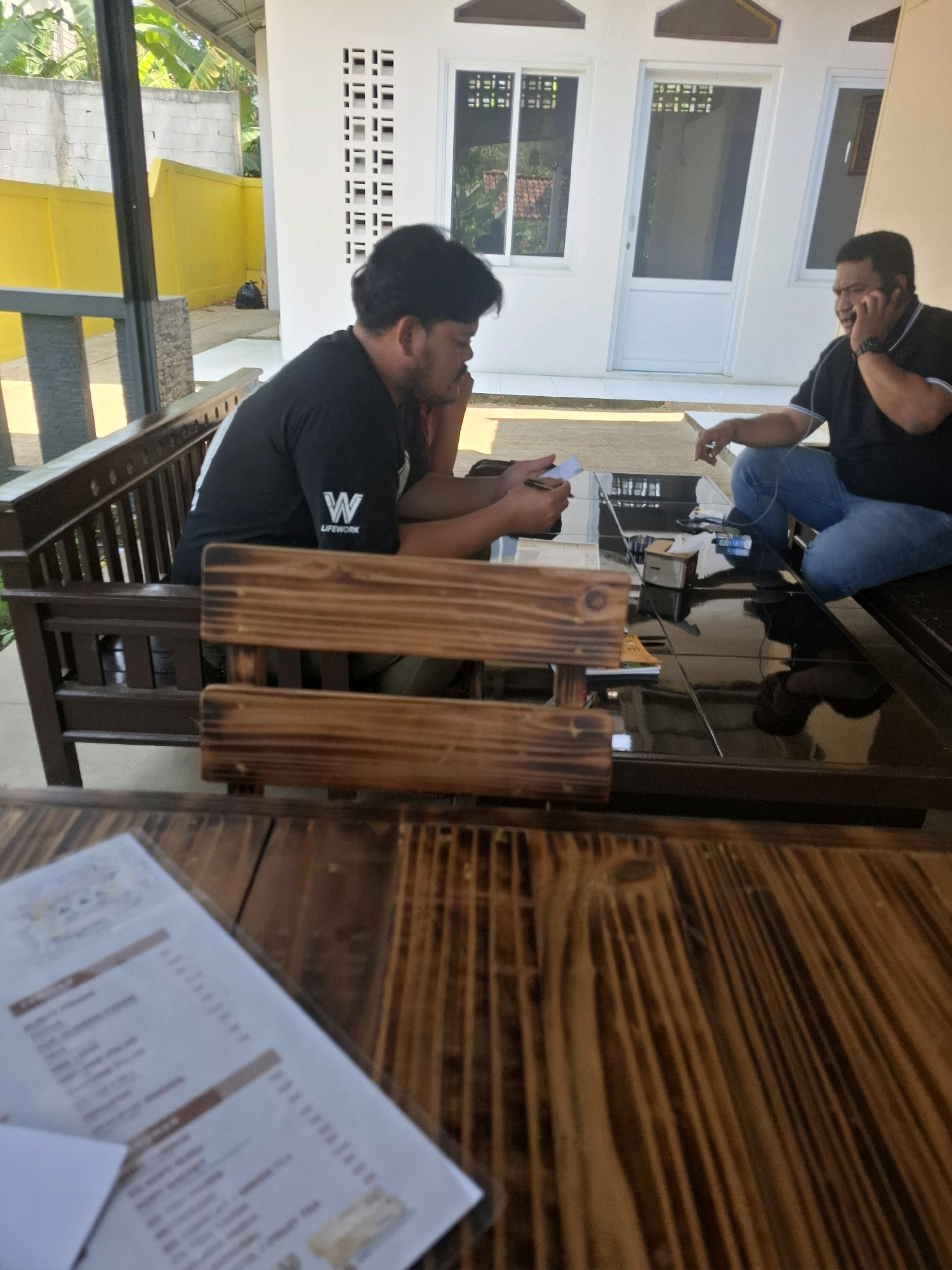 Two men are seated at a dark, reflective table outdoors. The man on the left, wearing a black t-shirt with a white "W LIFEWORK" logo, is looking down at a piece of paper and holding a pen. The man on the right, in a black polo shirt and blue jeans, is speaking on a mobile phone. They are positioned on a wooden bench, with another wooden bench and table in the foreground. The background features a white building with windows and doors, and a yellow wall. The scene appears to be in a casual outdoor setting, possibly a cafe or restaurant. The location is identified as Cibentang, Indonesia, suggesting a tropical climate given the presence of lush greenery and bright sunlight.