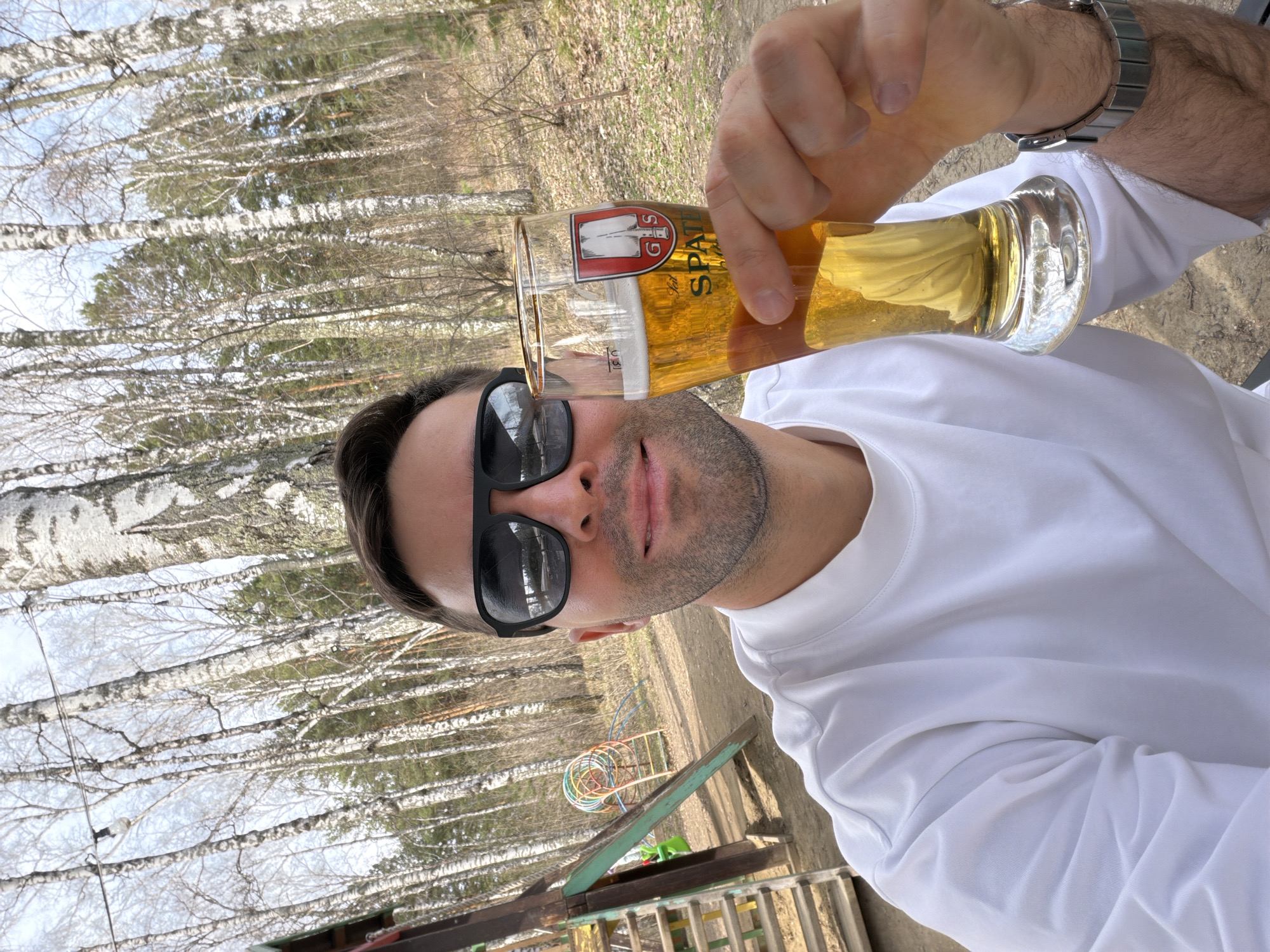 A fair-skinned man with dark hair and sunglasses is holding a glass of beer, likely Spaten beer, in an outdoor setting with a view of birch trees. He's wearing a white long-sleeved shirt and a wristwatch. In the background, there is a children's playground with a colorful, circular toy. The scene appears to be in a park or recreational area. The location might be within the city of Krasnoyarsk, Russia, given the prompt.
