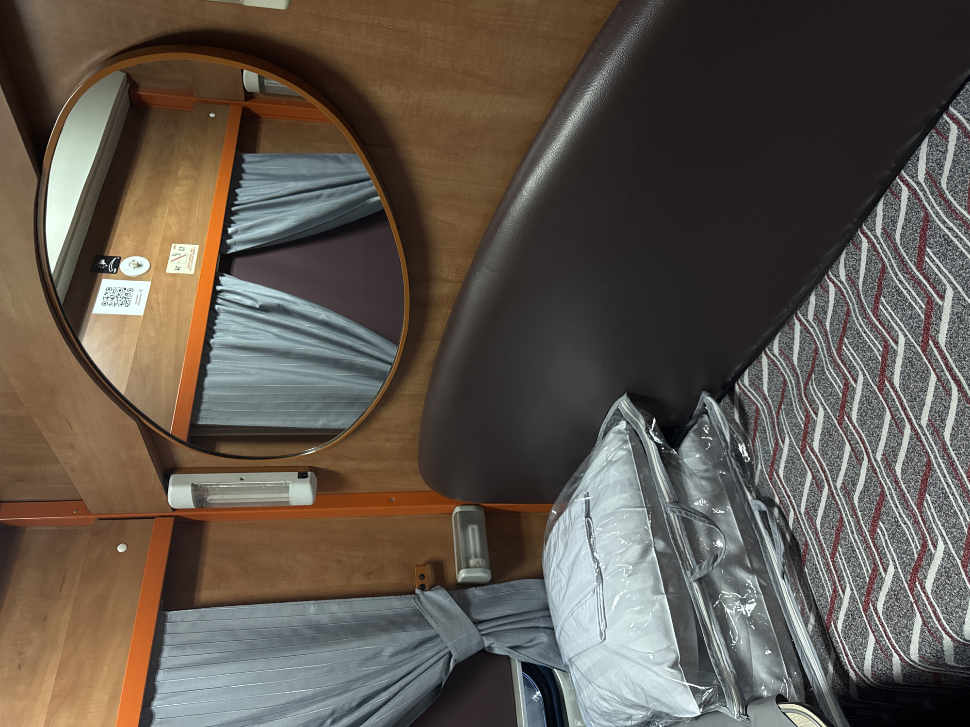 The image shows a small sleeping compartment, likely on a train, with wood-paneled walls and an orange metal frame. A dark brown upholstered seat or bunk runs along the right side of the compartment. On the left, light blue curtains are drawn. There are two stacked pillows in clear plastic packaging on the bed. A large oval mirror hangs on the back wall, reflecting the curtains and some stickers. A fluorescent light fixture is mounted on the wall to the left of the mirror. The bed is covered with a patterned blanket in shades of gray, white, and red. The overall impression is that of a compact and functional sleeping space. There is no indication of the city of Moscow, Russia in the image.