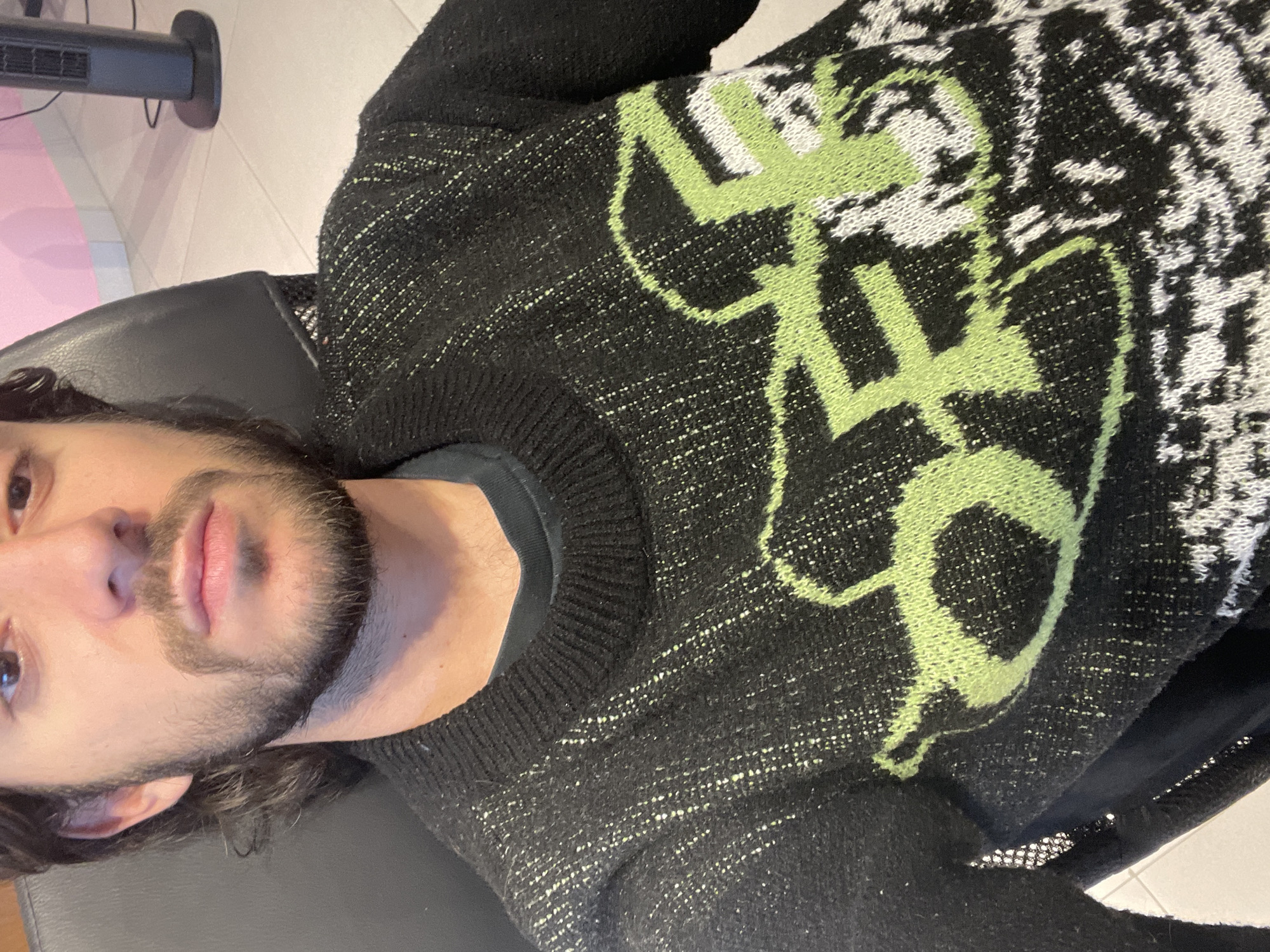 A man is lying on his back wearing a black and light green sweater with a black t-shirt underneath. He has dark hair and a beard. The background consists of a black chair, a light-colored floor, and a pink-colored wall. The image does not contain enough information to determine the location in Mexico City.
