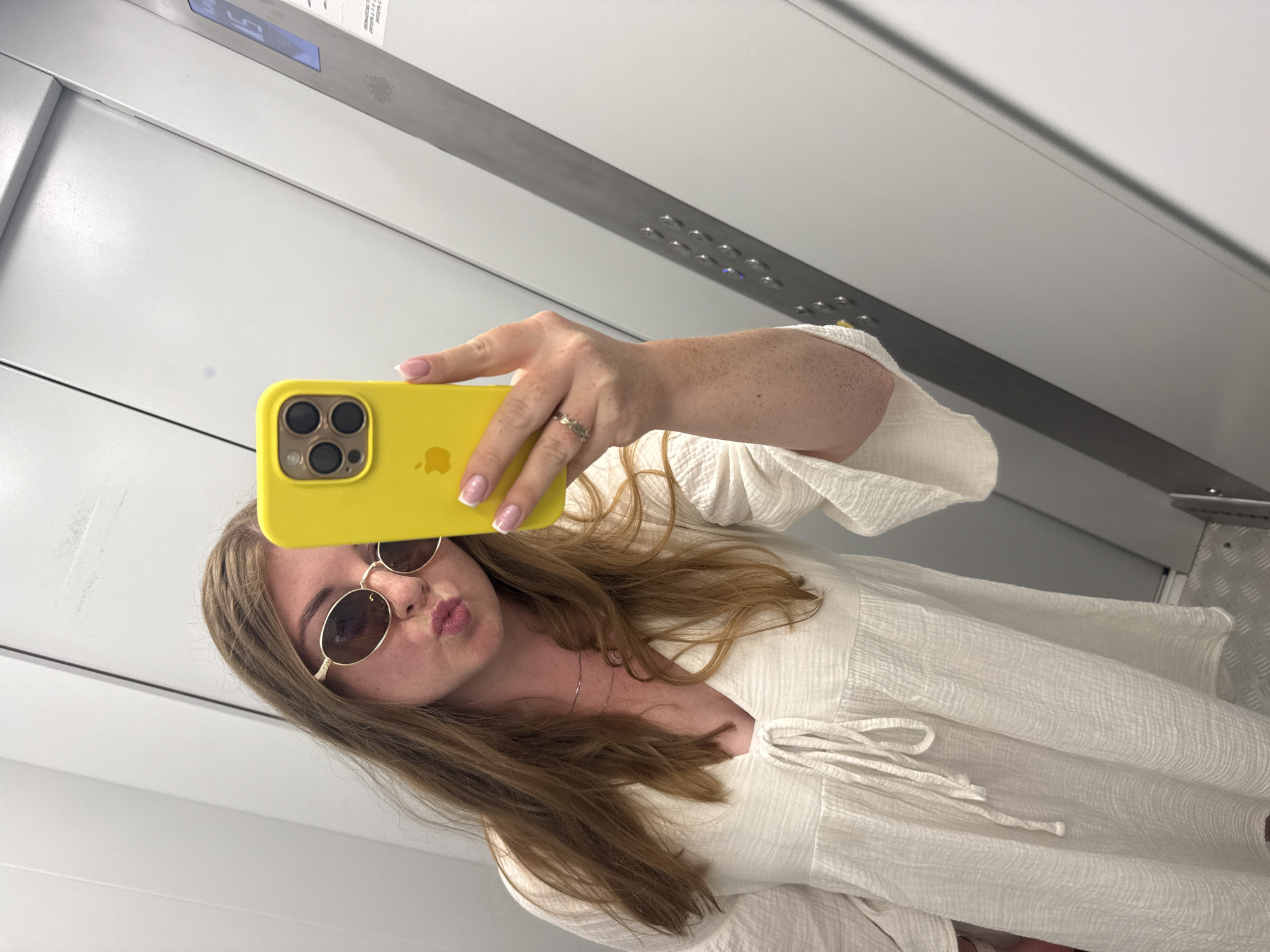 A woman is taking a selfie in an elevator. She is wearing a white dress, oval sunglasses, and a gold necklace. She is holding up a yellow iPhone to take the photo. The elevator is gray and has a control panel with buttons. The floor of the elevator is metal.