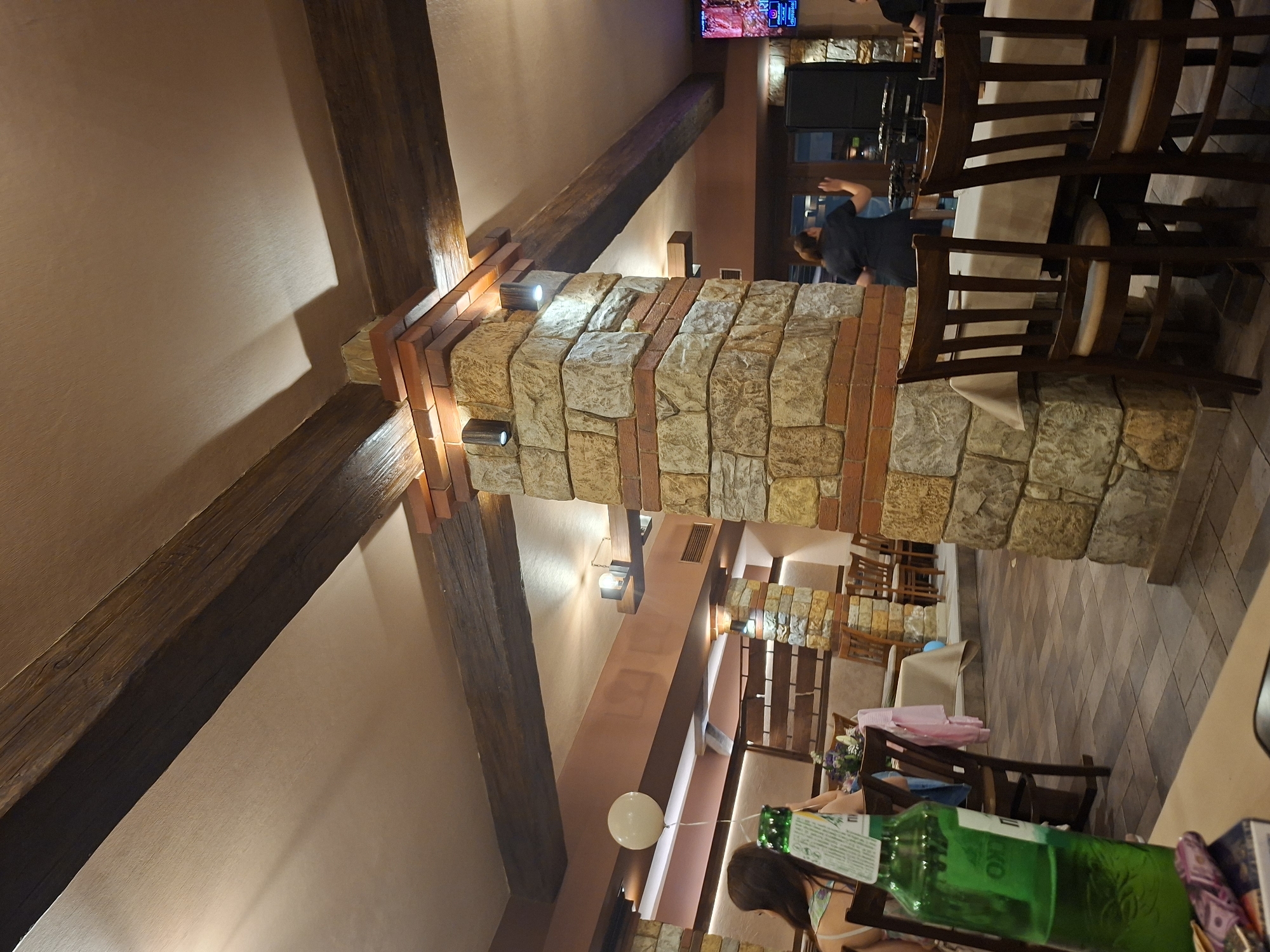 The image depicts the interior of a restaurant. The architecture features prominent wooden beams and a stone and brick column with integrated lighting. Tables and chairs are visible, some appearing to be set for dining. At least one person is visible in the background, possibly a waiter or staff member. A green bottle and other items are on a table in the foreground, indicating a recent or ongoing meal. The location appears to be in Pleven, Bulgaria based on visual elements.
