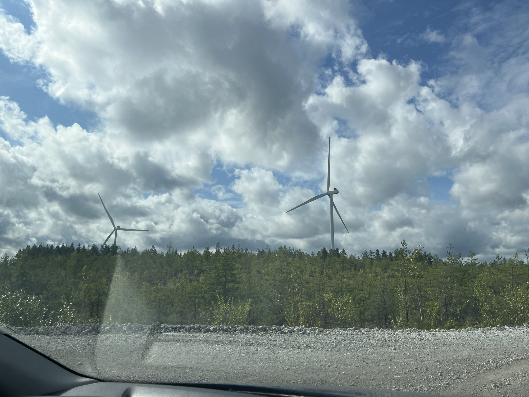 In Aidu, Estonia, a dirt road stretches across the foreground. Beyond the road, a dense line of green trees forms a backdrop. Two wind turbines stand tall against a partially cloudy sky. The sky is a mix of blue and white clouds, with some areas of darker, more ominous clouds visible. One wind turbine is closer to the left side of the image and appears to be slightly shorter than the one on the right. The wind turbine on the right is more centrally located in the frame and is in focus. The overall scene is one of a rural landscape with renewable energy infrastructure.
