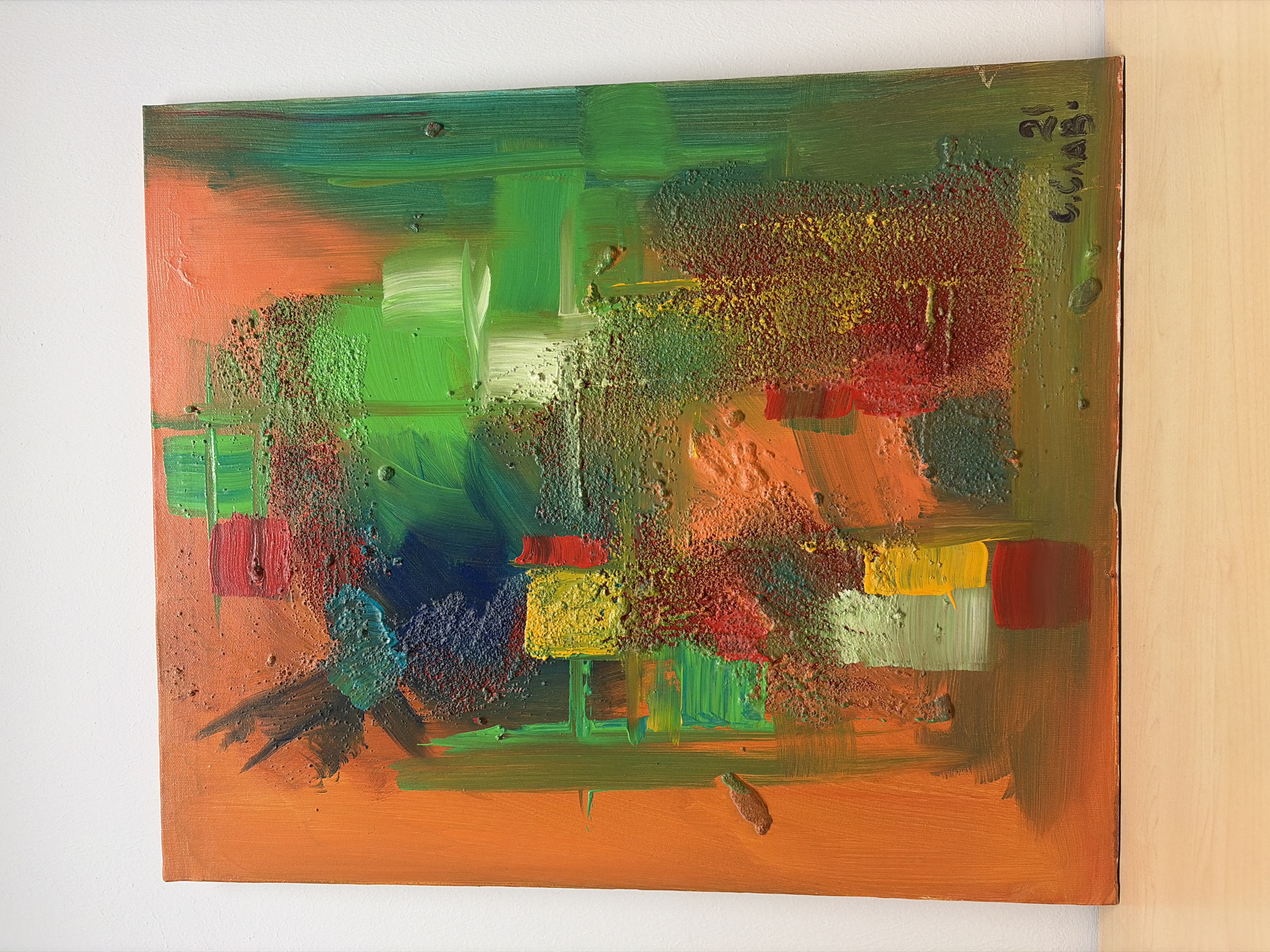 A framed, abstract painting dominates the scene. The painting is on a canvas and leans against two walls, one white and the other light brown. The artwork features a predominantly orange background layered with patches of green, red, yellow, and blue. The paint application varies, with some areas appearing smooth while others have a textured, almost granular quality. Brushstrokes are visible, and some areas have a thick impasto. On the upper right corner of the painting, there is a signature in dark ink. There is no indication this image was taken in Sofia, Bulgaria.