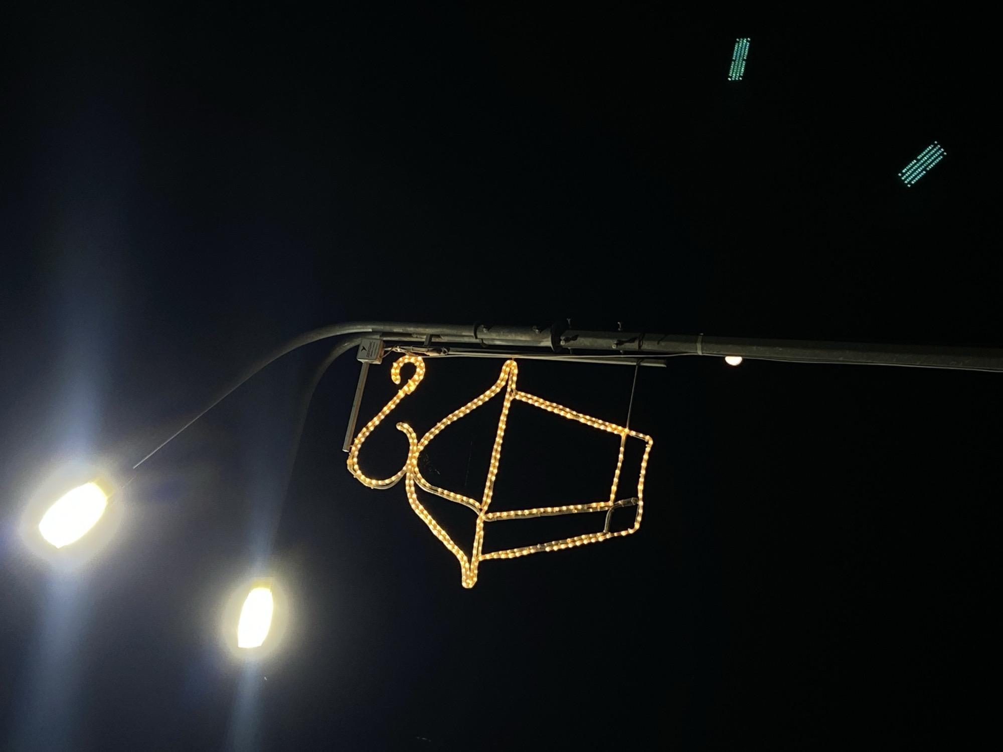 The image captures a nighttime scene in Tolyatti, Russia, focusing on a street light pole adorned with festive lighting. Two bright, oval-shaped street lights illuminate the upper portion of the frame against a dark sky. Suspended from the street light pole is a decorative, lantern-shaped outline made of yellow LED lights. This decorative element features a curved handle extending from the top and a hexagonal base. The pole itself is metallic and gray, with visible wiring and a small, rectangular label attached to it. In the background, at least two sets of green, horizontal lights are visible, suggesting other urban lighting or traffic signals in the distance. The overall environment appears to be a street or public area at night, characterized by artificial illumination against a dark backdrop.