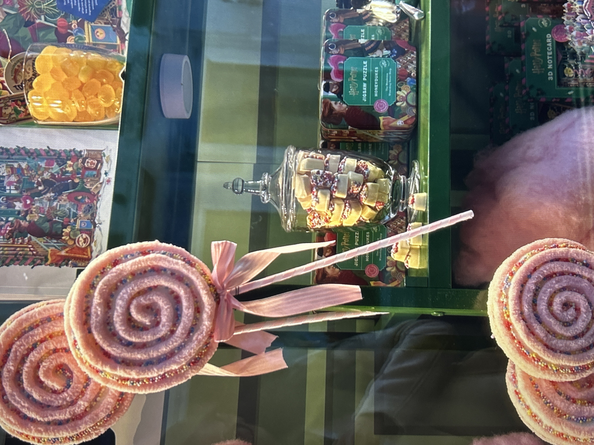 This image shows a display of sweets and toys likely found in a shop themed around the "Harry Potter" franchise. In the display, there are jars of candy, jigsaw puzzles labeled "Harry Potter," and "3D Notecards." Dominating the foreground are large pink swirl lollipops decorated with colorful sprinkles. A pink bow is tied to one of the lollipops. It's difficult to pinpoint the exact location in Gorna Oryahovitsa, Bulgaria, without additional context. The image does not portray people, nor does it imply particular actions or interactions. The environment is that of a store display, brightly lit and showcasing various merchandise.
