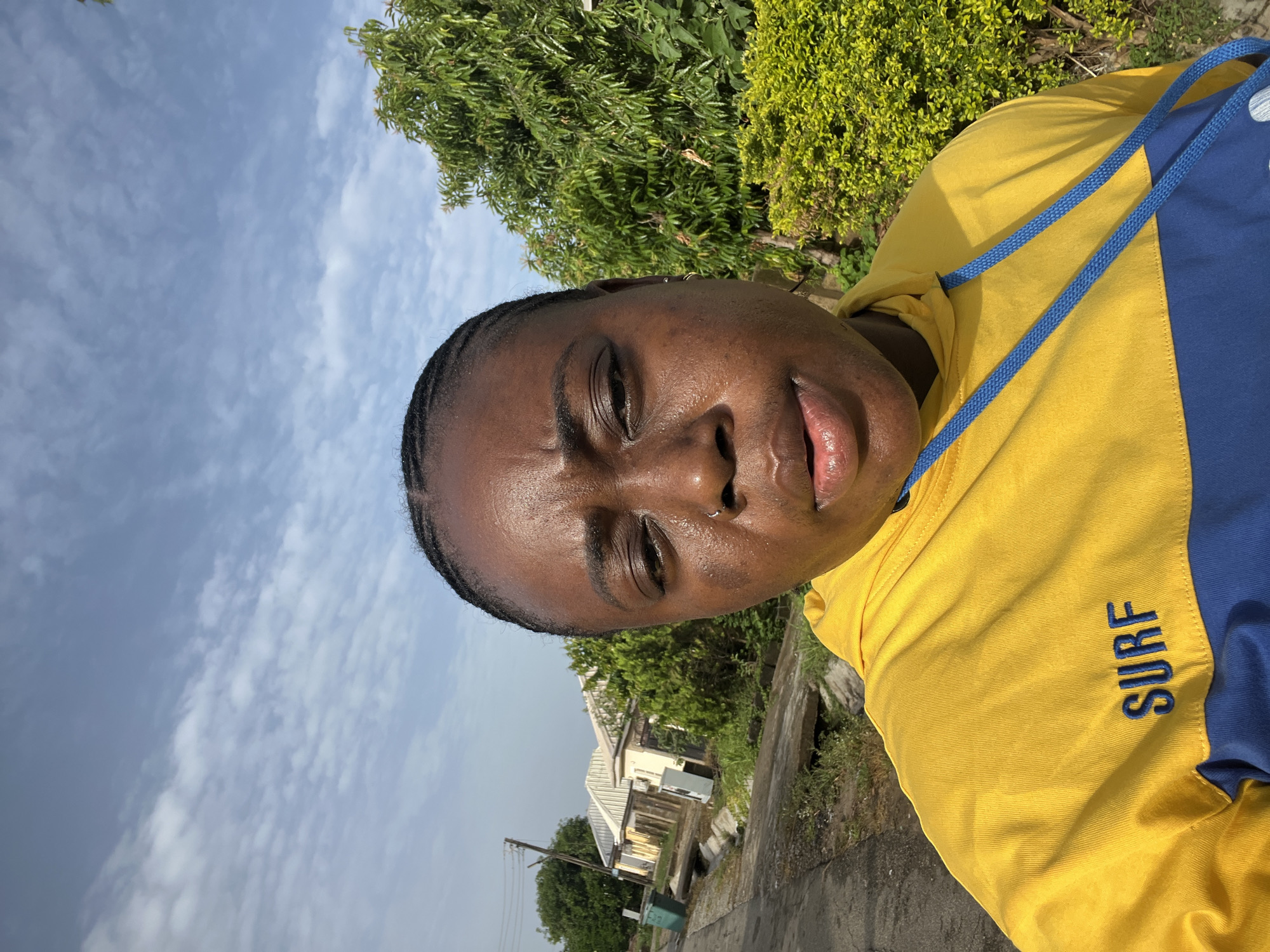 A close-up, slightly angled shot depicts a dark-skinned person with short, braided hair wearing a yellow and blue "SURF" hoodie with blue drawstrings. The person's face appears sweaty. The background shows a cloudy sky, greenery, a house, and a concrete path. It is not possible to identify the location of Abuja from the information provided.