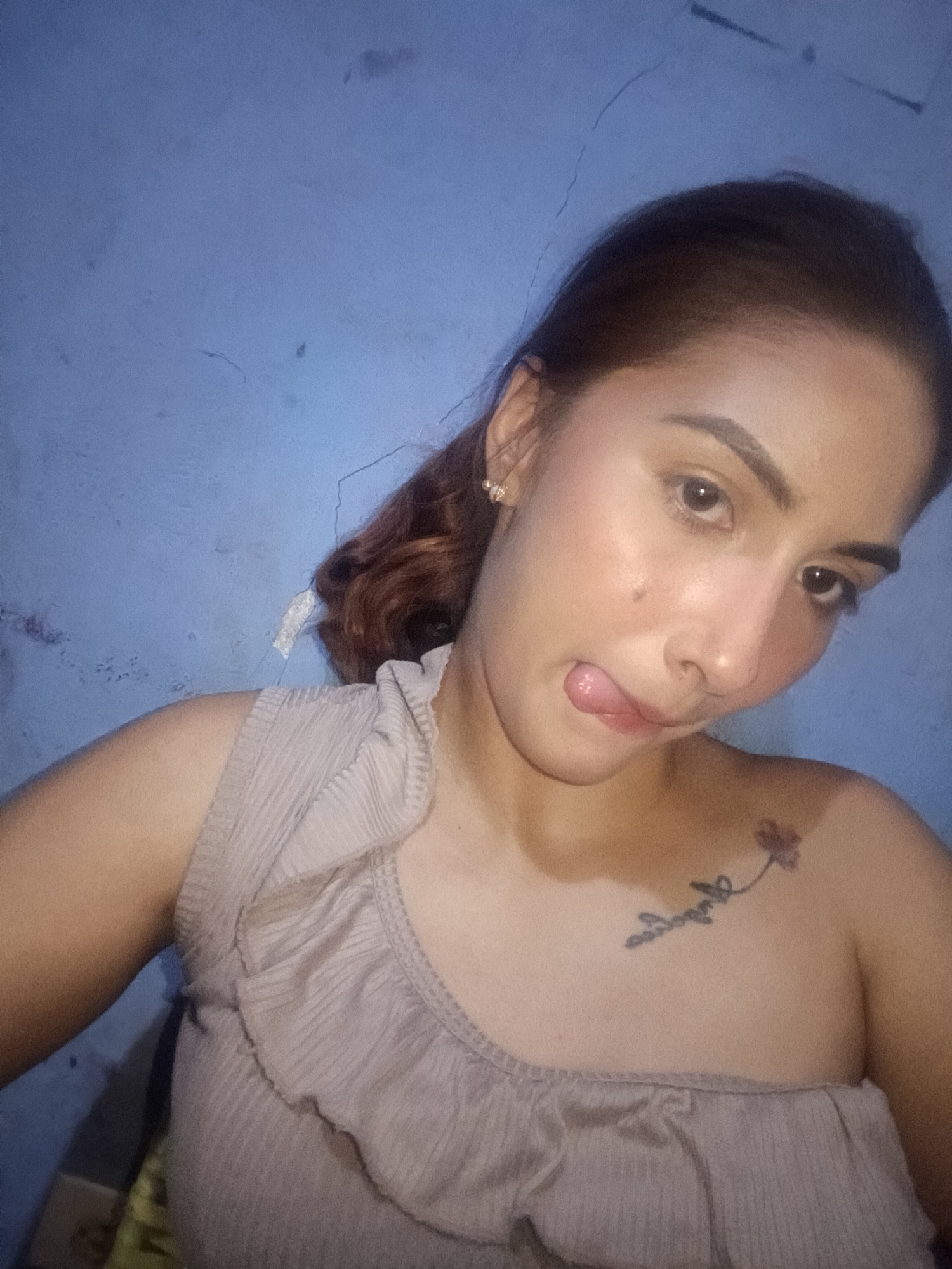 A woman is posing for a selfie. She has brown hair pulled back, light skin, and is wearing a one-shoulder gray top. She has a tattoo on her left collarbone and her tongue is sticking out. The background is a plain blue wall. The location is unknown and there is no indication that this is in Juan Griego, Venezuela.