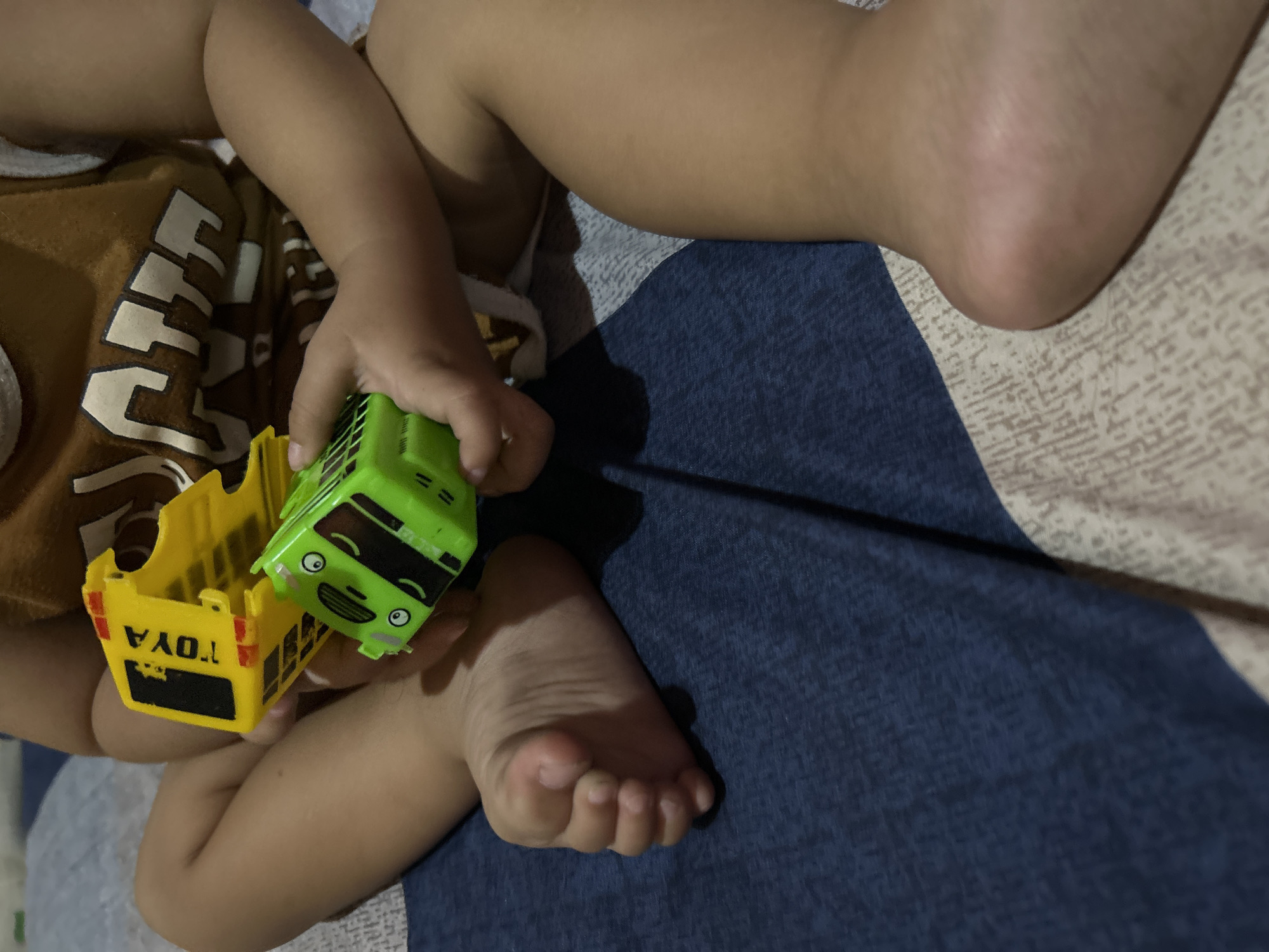 This media file shows a close-up of a child's legs and feet as they play with two toy vehicles. The child is wearing a brown t-shirt with a white graphic design. The toys appear to be a yellow truck and a green bus. The yellow truck has the word "TOYA" printed in black on its side. The child's hands are visible, holding the toys. The background consists of a surface with a patterned fabric, part of which is a dark blue color. The scene is indoors, and the lighting suggests it could be nighttime or a dimly lit room. The overall mood is one of playful engagement. The location context is Depok, Indonesia.