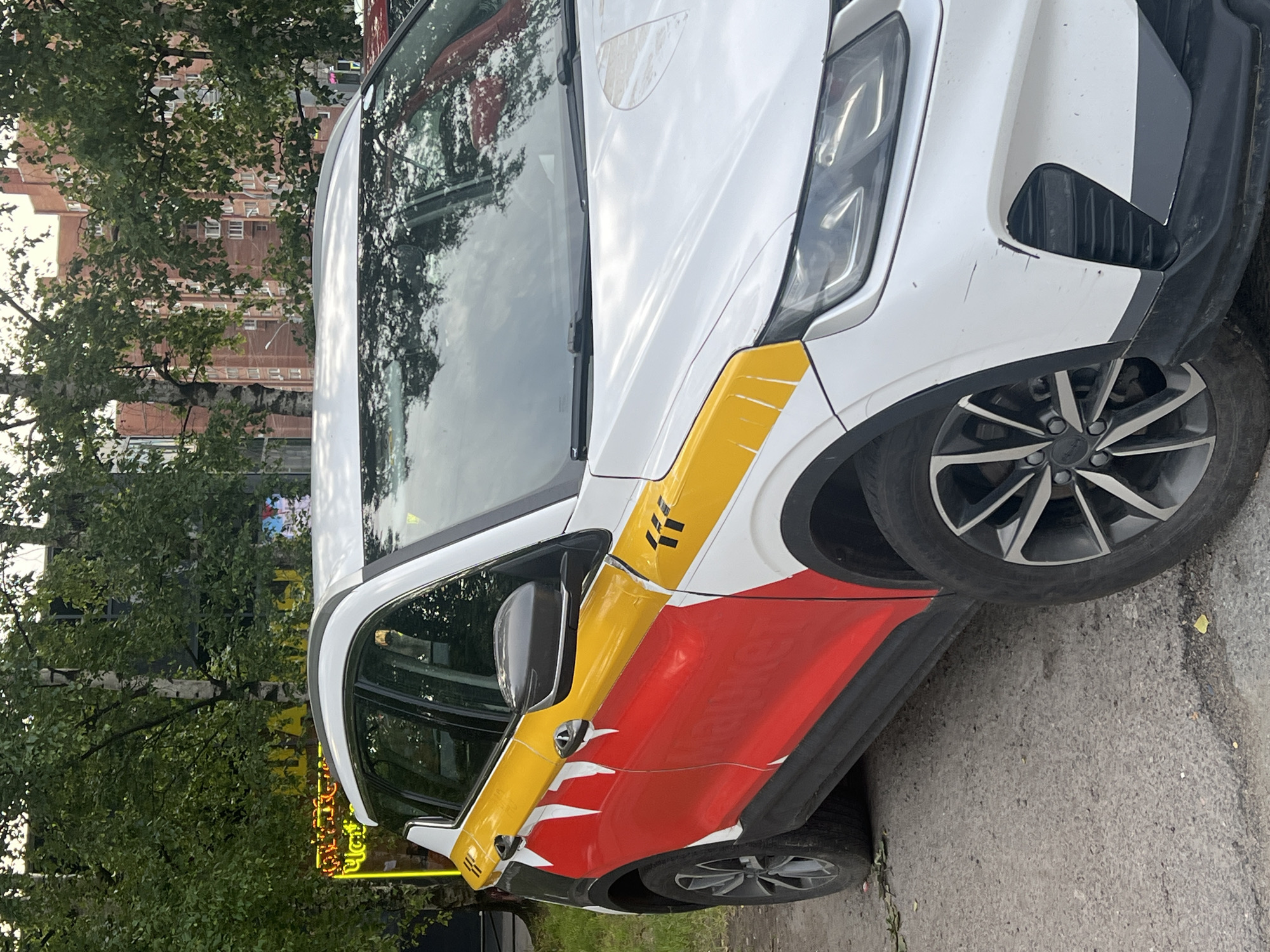 The image shows the side and front of a white car with a distinctive red and yellow livery, parked on a paved surface in front of trees and buildings. The car is a crossover SUV and has prominent yellow stripes along the top of the side panels, with black and red graphics on the doors. The front of the car features headlights and a scratched bumper. In the background, there are trees with green foliage and a multi-story brick building. A bright yellow neon sign is visible to the left of the car, behind the trees. The scene appears to be in an urban or suburban environment. Based on the visual cues, this image is likely located in Sinyavino, Russia. There are no people visible in the image.