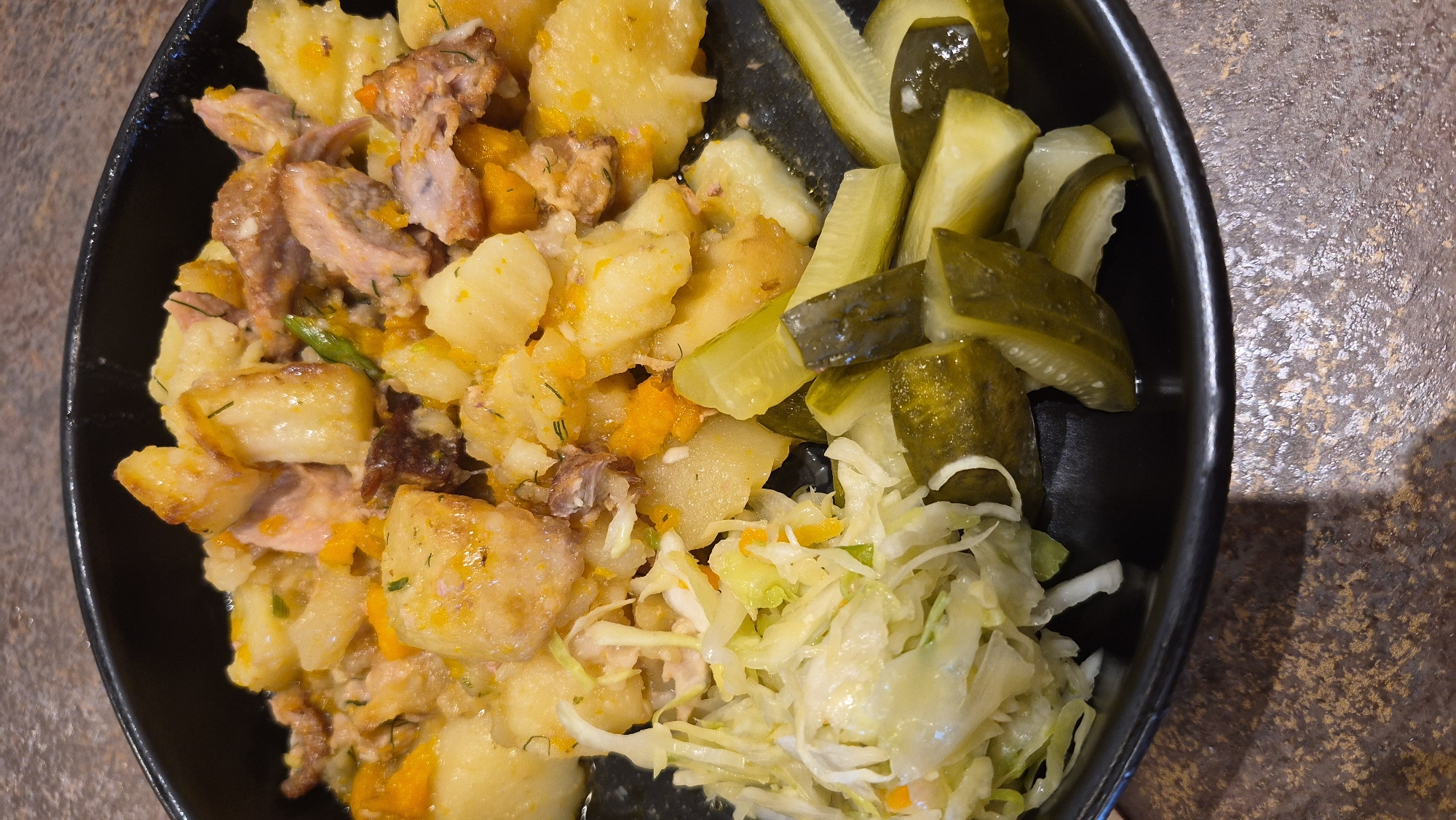 A close-up, top-down shot shows a black bowl filled with a hearty meal of potatoes, meat, and vegetables. The dish appears to be a stew or a roasted mixture. The potatoes are cut into bite-sized chunks and appear to be roasted, some showing crispy edges. Interspersed with the potatoes are pieces of tender meat, possibly pork or beef, with a reddish-brown hue and visible muscle fibers. Bright orange pieces of what look like carrots or sweet potatoes are also present, adding color to the dish. Scattered throughout the mixture are flecks of green herbs, likely dill or parsley, suggesting a savory seasoning.
To the left of the main dish, a generous portion of coleslaw is piled. The coleslaw consists of finely shredded white cabbage and some shredded carrots, mixed with a light dressing, giving it a fresh, crisp appearance.
On the right side of the bowl, several dill pickle spears are artfully arranged. The pickles are a vibrant green, with visible ridges and seeds, indicating they are likely brined or fermented.
The food is presented in a dark, possibly black, ceramic bowl with a slightly matte finish. The bowl rests on a textured surface that appears to be a tabletop with a mottled brown and gray pattern, resembling a speckled granite or laminate.
The lighting in the image is natural, casting subtle shadows and highlighting the textures of the food and the surface it rests on. The focus is sharp on the food, with the background slightly blurred.
The scene appears to be an indoor setting, likely a restaurant or a home dining area. There are no people or animals visible in the frame. The overall impression is one of a wholesome, satisfying meal.
Regarding the location in Daugavpils, Latvia, this specific dish, with its combination of potatoes, meat, coleslaw, and pickles, is not uniquely tied to any particular landmark or area within Daugavpils. It represents a common type of comfort food that could be found in various eateries or homes throughout the city. Without additional context, pinpointing an exact location within Daugavpils is not possible based solely on the visual information provided.