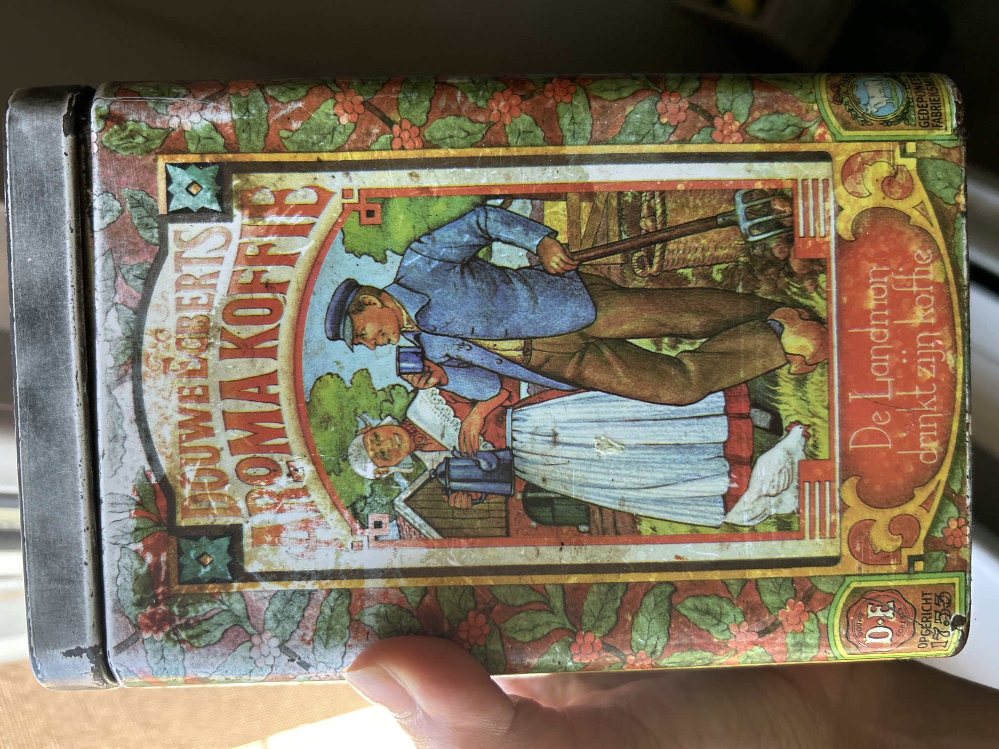 The image shows a rectangular tin can, likely for coffee storage, featuring a pictorial scene. A farmer in blue attire and cap is depicted with a pitchfork, while an older woman in traditional dress stands beside him, holding a container. A white chicken is visible in the background, near a building. The text on the can includes "DOUWE EGBERTS ROMA KOFFIE" and "De Landman drinkt zijn koffie." The can is held by a hand. The text on the bottom reads "OPGERICHT 1753". The top right corner also contains text. The image does not show any visual elements related to Kadievo, Bulgaria.