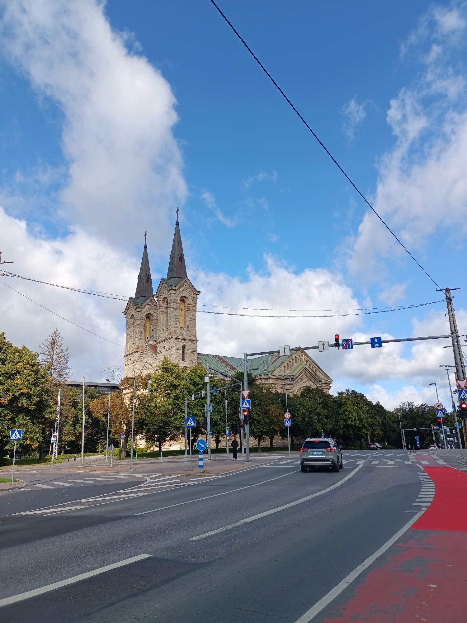 The image shows a street scene in Tallinn, Estonia. In the background, the distinctive twin spires of the St. Charles' Church are visible. The church is constructed of light-colored stone and has a green roof. It is surrounded by lush green trees, some of which are showing signs of autumn foliage.
The foreground features a multi-lane road with pedestrian crossings and traffic lights. Several cars are present on the road, including a silver SUV in the right lane. Pedestrian crossing signs are visible on poles at the side of the road. The sky is a bright blue with scattered white clouds. Overhead power lines are also visible, stretching across the frame. The road surface is asphalt, and a portion of the right lane is marked with a red color, indicating a bike lane or a special traffic zone. The overall impression is a typical urban scene in Tallinn on a clear day.