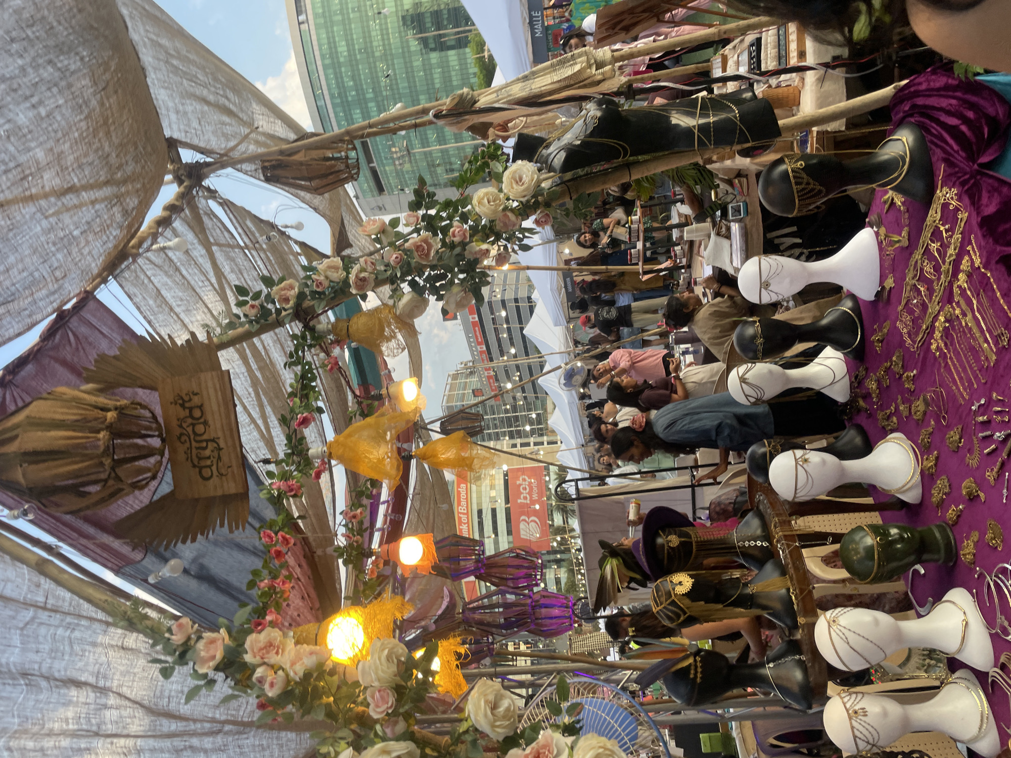 This image shows a vendor stall at an outdoor market in Mumbai, India. The stall is covered with a burlap roof adorned with flowers and decorative lights. A wooden sign with "drydaa?" is hung on the left side. The display table is covered with a purple cloth showcasing various fashion accessories displayed on mannequin heads of different colors, including gold jewelry. People can be seen browsing the merchandise. Behind the market stall are high-rise buildings with advertisements for "Bank of Baroda" and "bob World," indicating the location is likely within a commercial district of Mumbai.