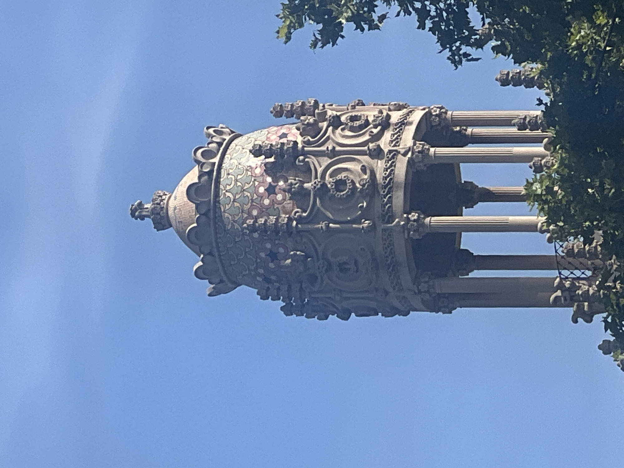 The image shows a detailed view of a decorative turret or pinnacle against a clear blue sky. The structure is ornate, with a domed top adorned with colorful mosaic tiles in a scale-like pattern of pink, white, and green. Below the dome, the turret features elaborate stone carvings, including circular motifs and what appear to be grotesque faces or gargoyles. The turret is supported by a colonnade of slender columns. To the right, the lush green foliage of a tree partially obscures the view of the structure, with its leaves creating a dappled effect against the sky and architecture. The style of the architecture suggests it is located in Barcelona, Spain, possibly a part of a modernist building such as Park Güell or another Antoni Gaudí designed structure. There are no people visible in the image.