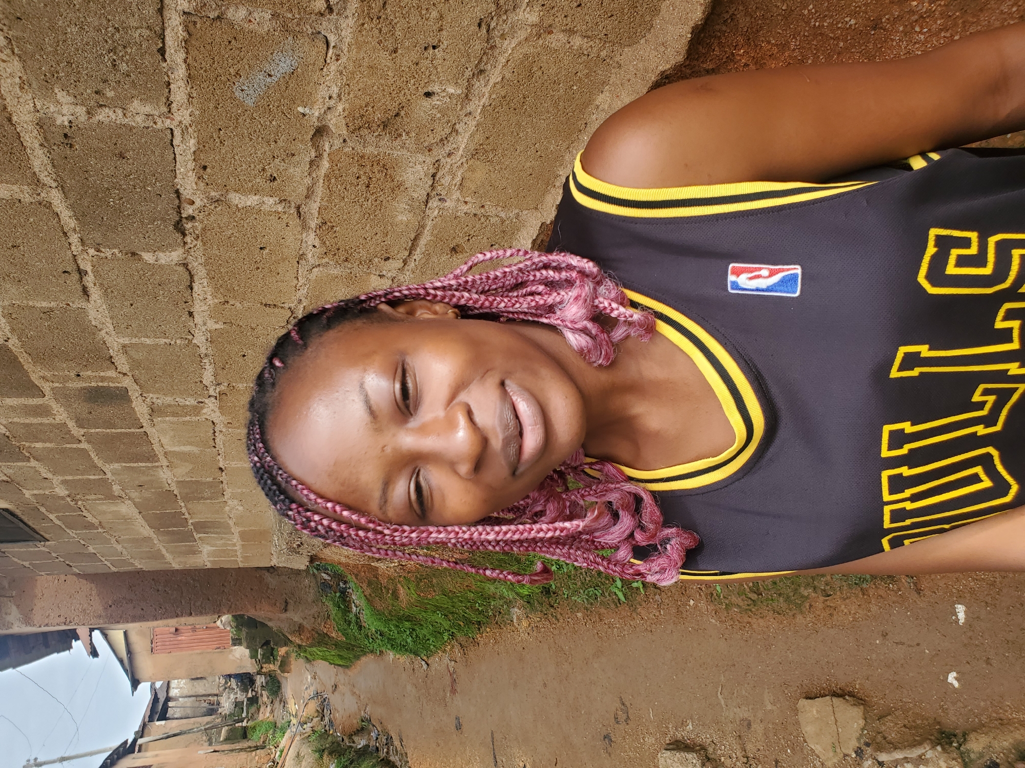The image shows a young woman with pink braided hair, wearing a black basketball jersey with yellow trim and the NBA logo. She is lying down, resting her head on a textured brick wall. Her eyes are closed and she has a gentle smile on her face, suggesting a relaxed or content emotion. The background shows a somewhat informal setting with glimpses of what appear to be homes or structures built with brick and corrugated metal, along with some greenery and a dirt path. This scene is consistent with a residential area in Jos, Nigeria. The lighting suggests it is daytime. There is yellow text visible on the jersey, but it is partially obscured and difficult to read definitively.