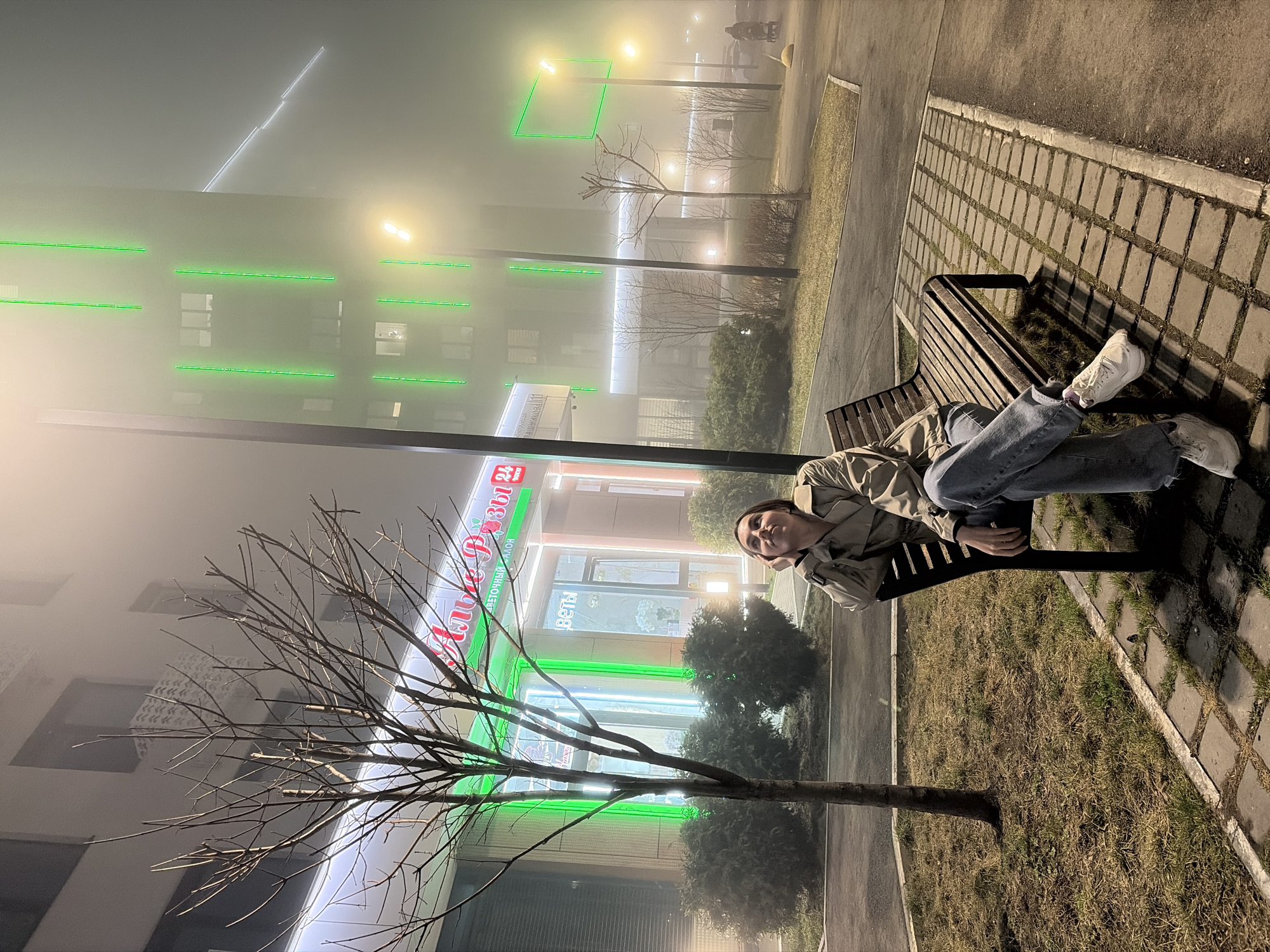 A person reclines on a bench in a foggy urban scene. The person is wearing light blue jeans, white sneakers, and a tan jacket. They are resting their head on their hand, facing the viewer.
Behind the bench is a paved area with grass, and a tree with bare branches in the foreground. Further back is a building lit with green neon lines. A sign with Russian lettering and a "24" logo is visible on the building. The city appears to be Izhevsk, Russia, based on the Cyrillic lettering of the sign and the architectural style.
The scene is illuminated by streetlights, but the fog reduces visibility. The general setting is a park or public space in an urban environment.