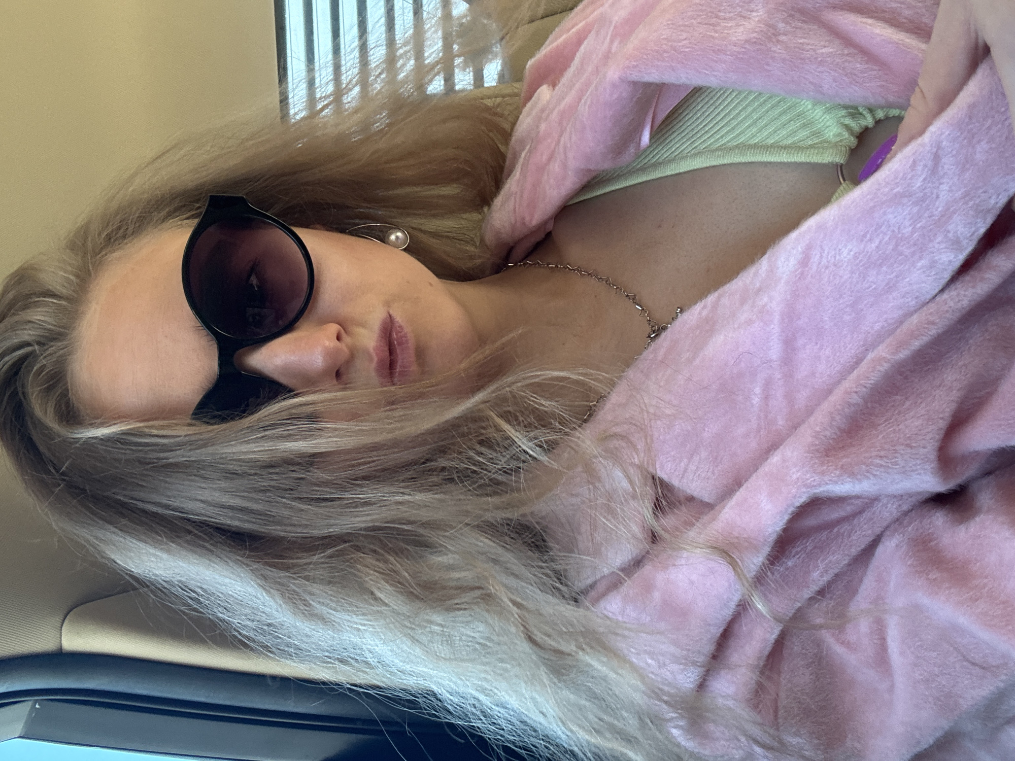 A blonde woman wearing sunglasses, a green top, and a pink jacket is sitting in a car. The location is Unknown, Unknown. She is also wearing a necklace and earrings. There are blinds visible in the background.