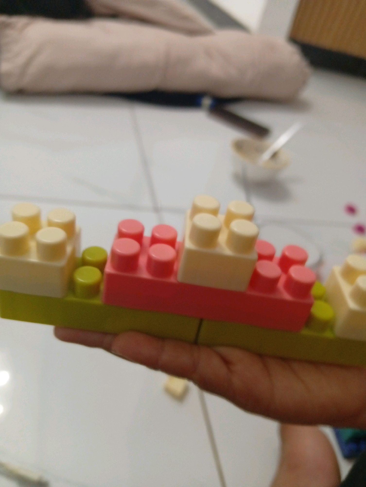 The image shows a close-up view of a child's hand holding a construction toy made of interlocking plastic blocks. The blocks are arranged in a layered structure, with a lime green base, a pink middle section, and ivory-colored blocks on top. There are also two small, bright yellow blocks visible between the ivory and pink sections. The background is out of focus and appears to be an indoor setting, possibly a home, with a patterned floor and a hint of furniture. The lighting suggests an indoor environment. The image does not provide any information or visual cues that would allow for the identification of its location in Samarinda, Indonesia, or any specific environmental context related to that city.
