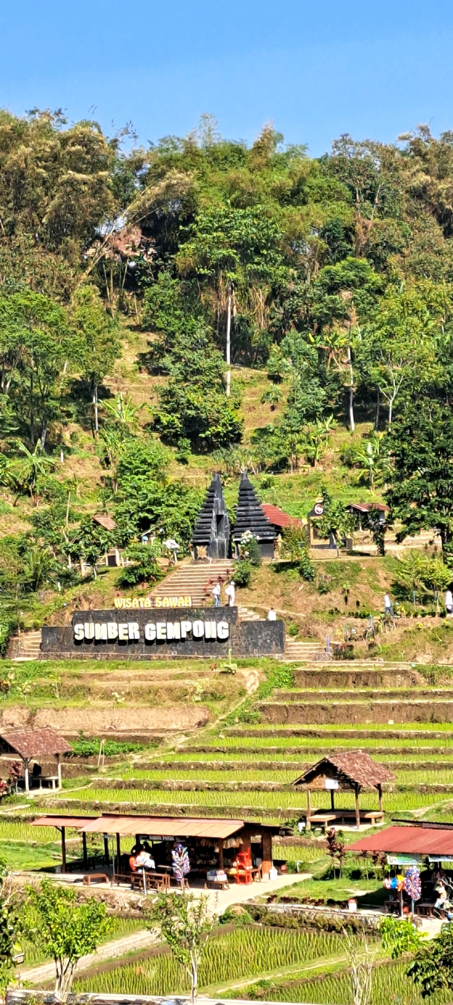 The image depicts the Wisata Sawah Sumber Gempong attraction in Tretes, Indonesia, showcasing a terraced rice paddy landscape under a clear blue sky. In the mid-ground, a large sign with raised white letters reads "SUMBER GEMPONG" above a smaller sign that says "WISATA SAWAH." Stone steps lead up to a structure with two dark, pyramidal towers. Several individuals are visible on and around the steps, some appearing to be tourists. The foreground and middle ground are dominated by lush green rice terraces, with small thatched-roof huts and open-air food stalls or shops scattered throughout. One stall in the lower right displays colorful merchandise. The background is a dense hillside covered with trees.