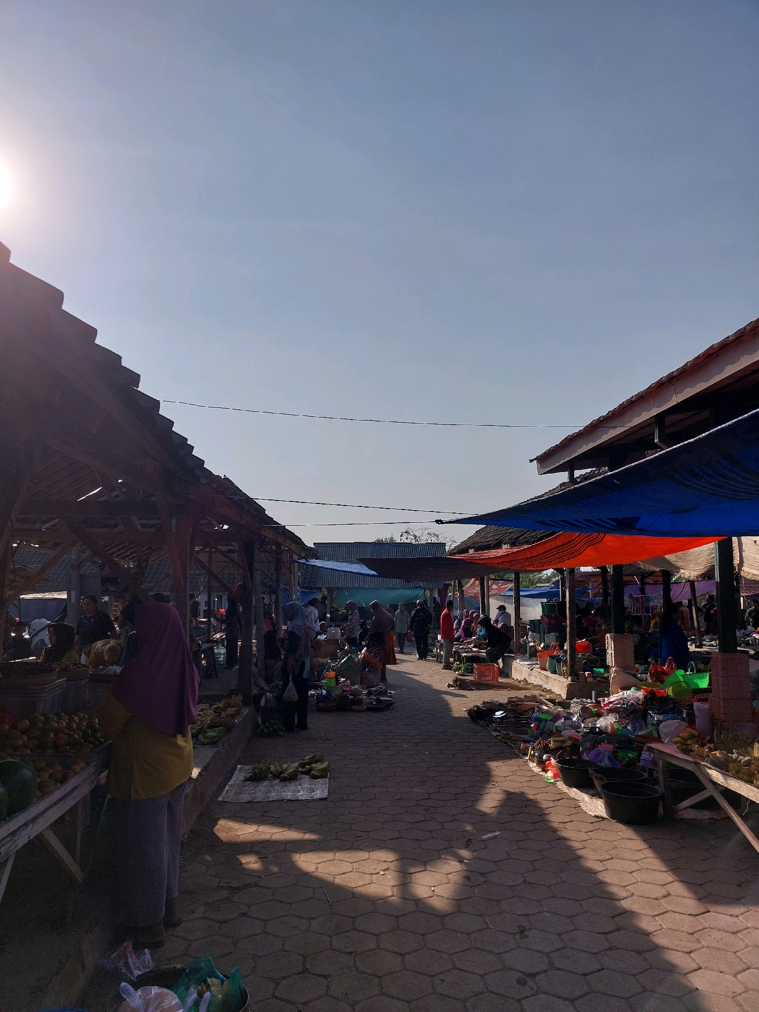 A bustling outdoor market is captured under a clear, bright sky. The sun is high and to the left, casting long shadows across the paved walkway. Stalls are set up along both sides of the path, covered with wooden roofs and awnings made of various materials, including blue and red tarps. The market appears to be in a tropical or subtropical climate, given the presence of fresh produce like fruits and vegetables displayed on tables and mats. Numerous people are present, browsing, selling, and interacting within the crowded market aisles. The overall atmosphere suggests a lively and active commercial hub. The image depicts an open-air market likely located in Sidorejo, Indonesia, characterized by its vibrant trade and community gathering.