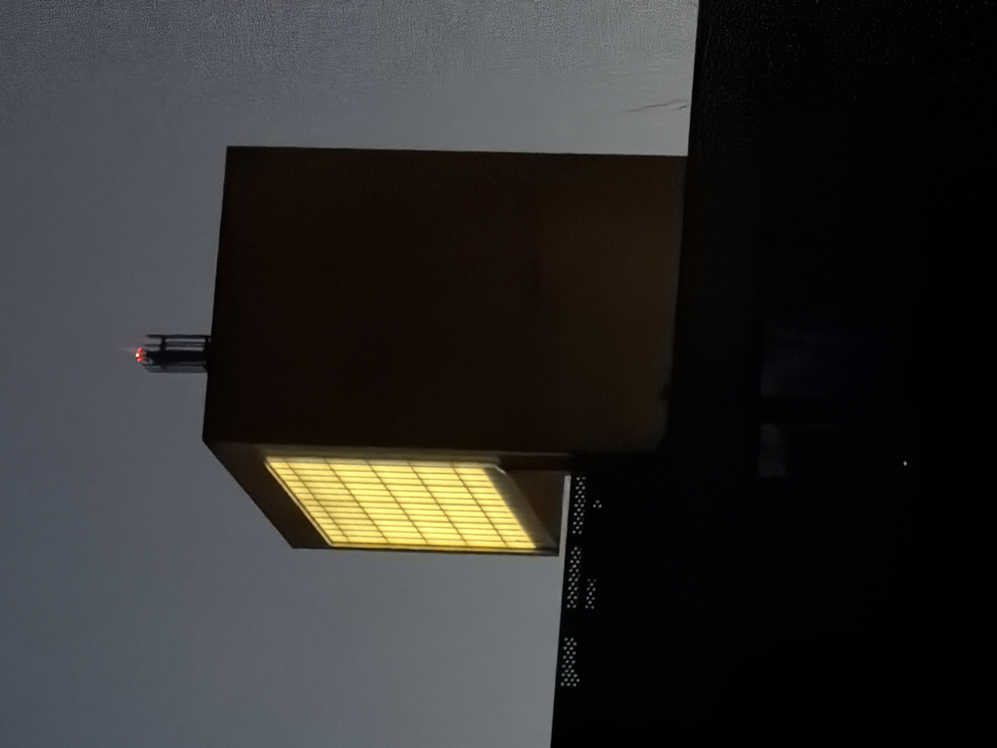 The image shows the top portion of a yellow rectangular building against a dark, overcast sky. The building has a large, illuminated window composed of many vertical rectangular panels, glowing with a warm yellow light. On the very top of the building, a black antenna-like structure is visible with two small red lights at its peak. In the foreground, a dark, silhouetted structure with a dotted pattern runs horizontally across the bottom of the image, partially obscuring the base of the building. The overall impression is of an industrial or commercial building seen at dusk or night. This building is located in Leudelange, Luxembourg.
