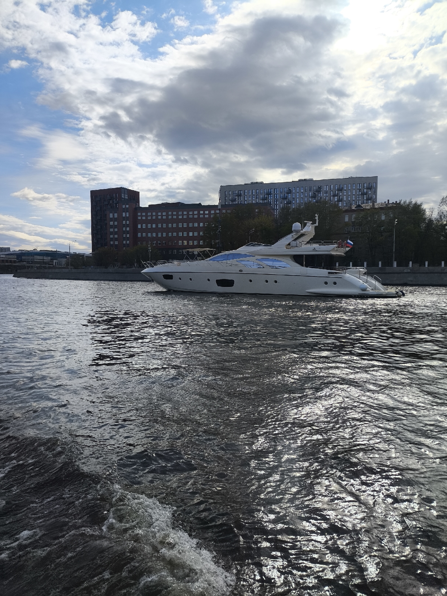 A white yacht is moving on the Moscow River. The Russian flag is visible on the stern. Buildings line the shore behind the yacht. The sky is overcast with mixed blue and gray clouds. The water is choppy with reflected sunlight.
