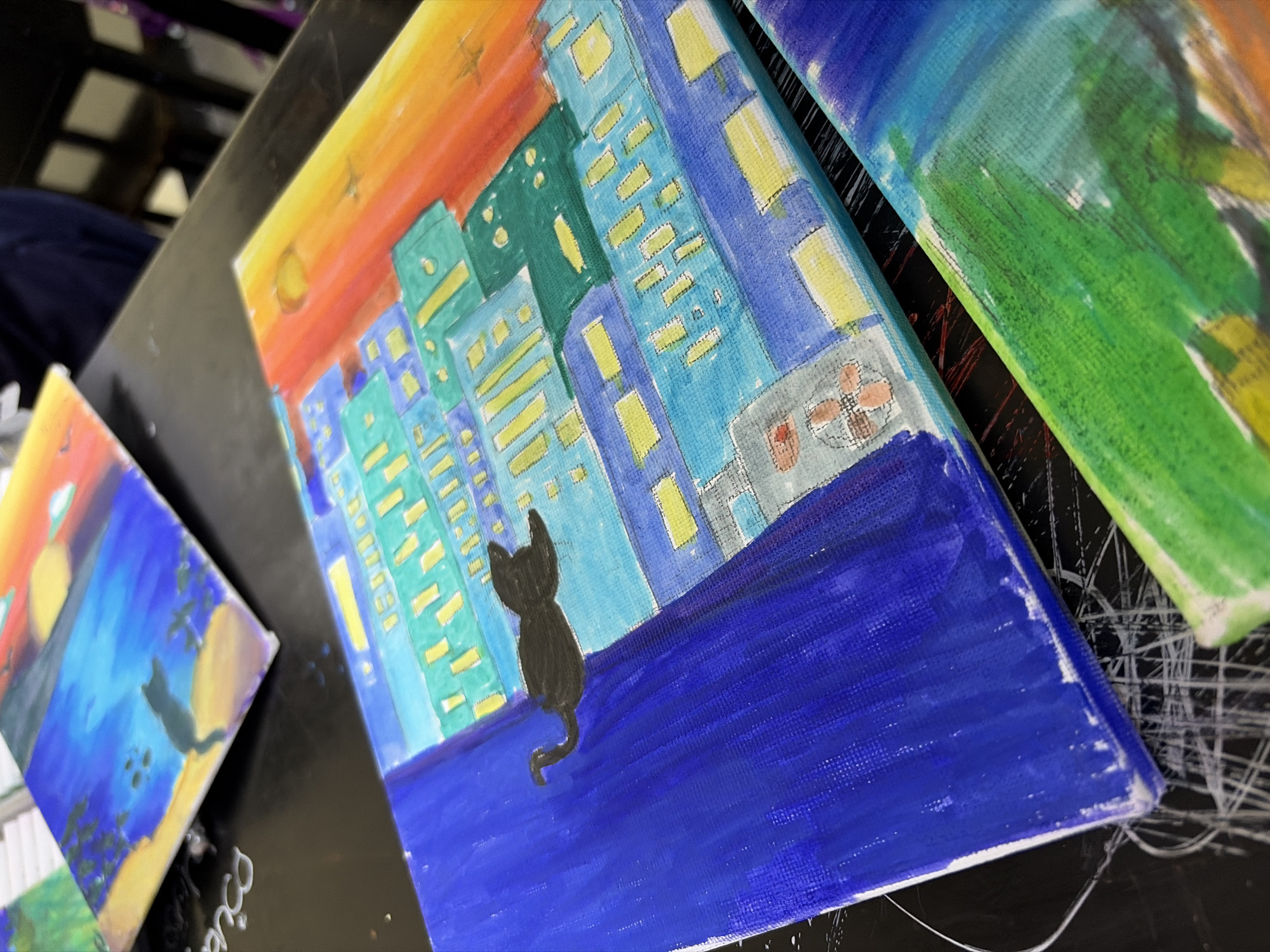 The image shows three small, painted canvases resting on a dark, reflective surface. The canvas in the center appears to depict a stylized cityscape at sunset or sunrise, featuring a black cat silhouette overlooking the scene from a dark blue ledge. The buildings are rendered in shades of blue and green, with yellow window details. The sky transitions from orange to yellow at the top of the canvas. Another similar painting with the cat in a slightly different pose is visible on the left. A third canvas, featuring primarily green hues, is partially visible on the right. The background appears blurry and out of focus. There is no indication this is Guayana City, Venezuela.