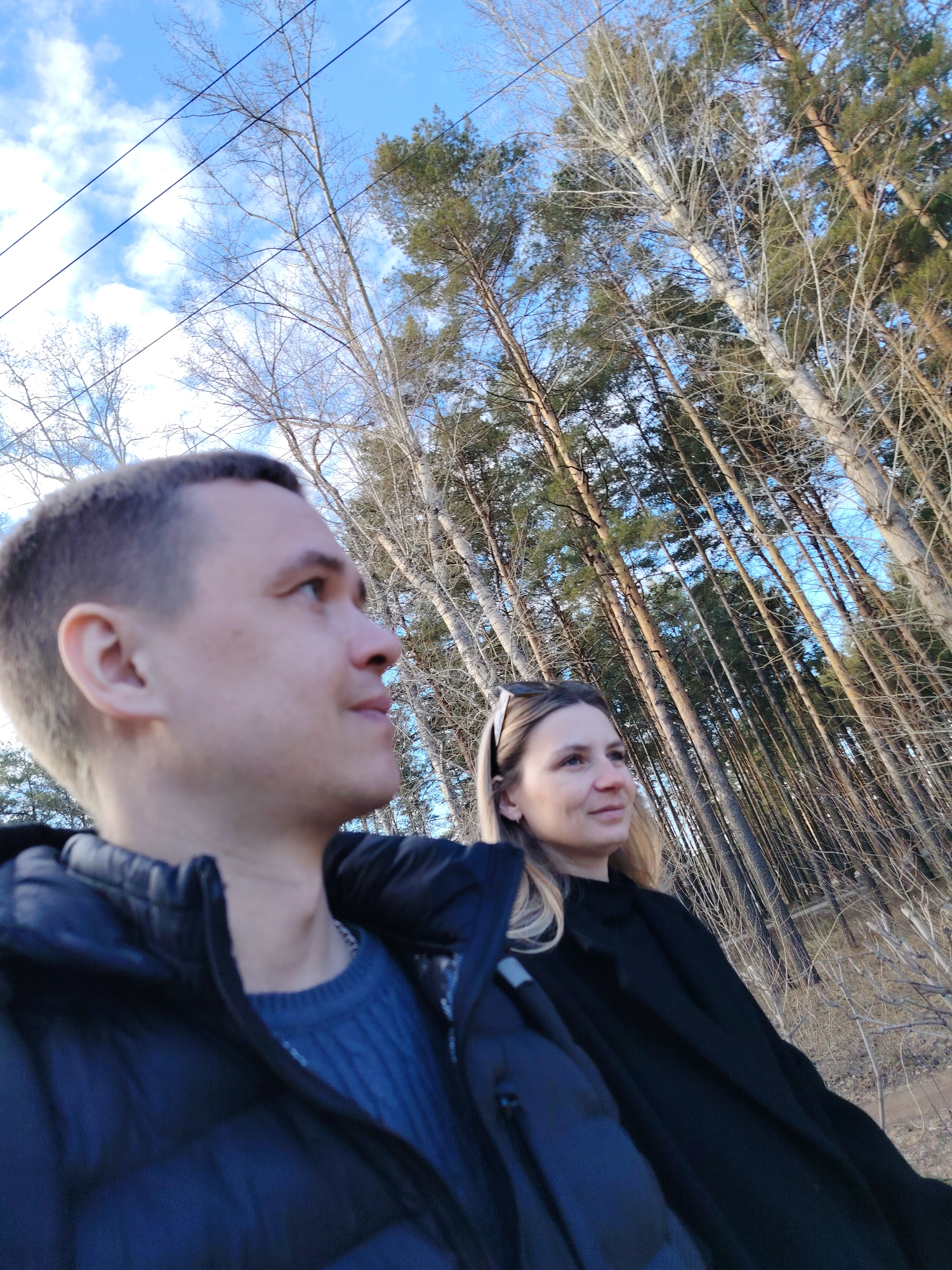 A photograph taken in Chaikovsky, Russia, shows two individuals, a man and a woman, standing outdoors in what appears to be a wooded area on a sunny day. The man is on the left, wearing a navy blue puffer jacket over a dark blue sweater. The woman, standing beside him, is wearing a black coat and has blonde hair with sunglasses perched atop her head. Both are looking off in the same direction to the right of the frame. The background consists of tall trees, some bare of leaves, others with dense green foliage, and a partly cloudy sky. Power lines are visible in the upper left corner of the image. The ground is covered in dry vegetation and branches.