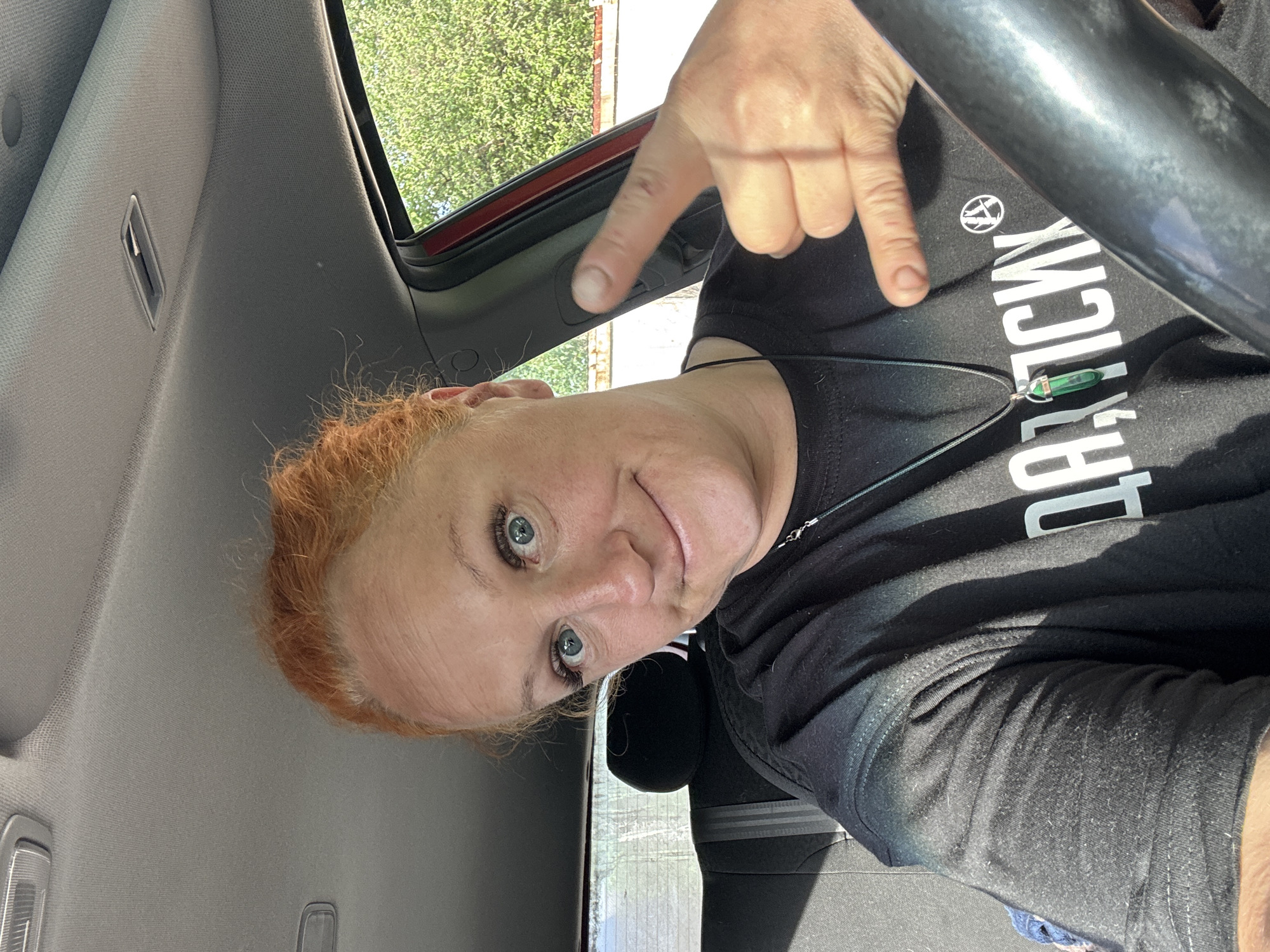 The image is an upward-angled selfie of a fair-skinned woman with short, auburn hair, light-blue eyes, and visible wrinkles. She is wearing a black T-shirt with white Cyrillic lettering and a silver necklace with a green pendant. Her right hand is raised with the index and pinky fingers extended, forming a "rock on" hand gesture. She is inside a car, with the steering wheel visible at the top right. The interior of the car is gray, and the outside is a green leafy tree. The lettering on the shirt seems to say "Да! ТЫСЯЧИ". The location could not be verified as Chaikovsky, Russia.