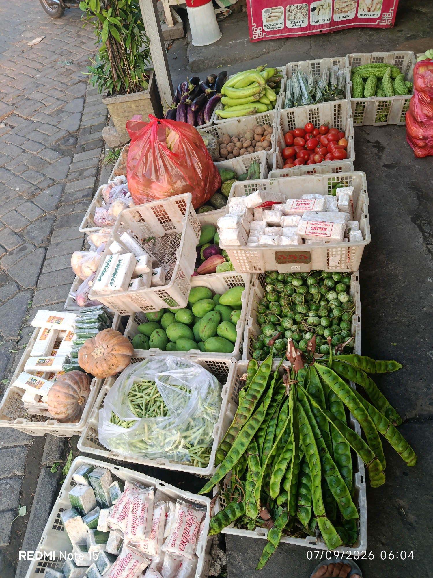 The image shows an outdoor market stall in Keboansikep, Indonesia, with a variety of fresh produce displayed in white plastic crates on a cobblestone ground. The stall features eggplants, long green beans, bitter gourds, tomatoes, potatoes, mangoes, small green eggplants, pumpkins, and bundles of petai beans. Packaged tempeh is also visible in several crates. A red plastic bag filled with produce sits on the ground next to some crates. A portion of a plant in a pot and parts of a motorcycle are visible on the left side of the image. A sign with Indonesian text and images of food items is displayed at the top of the stall. The timestamp "07/09/2026 06:04" and "REDMI Note 15" are present in the bottom right and left corners respectively. The lighting suggests it is daytime.