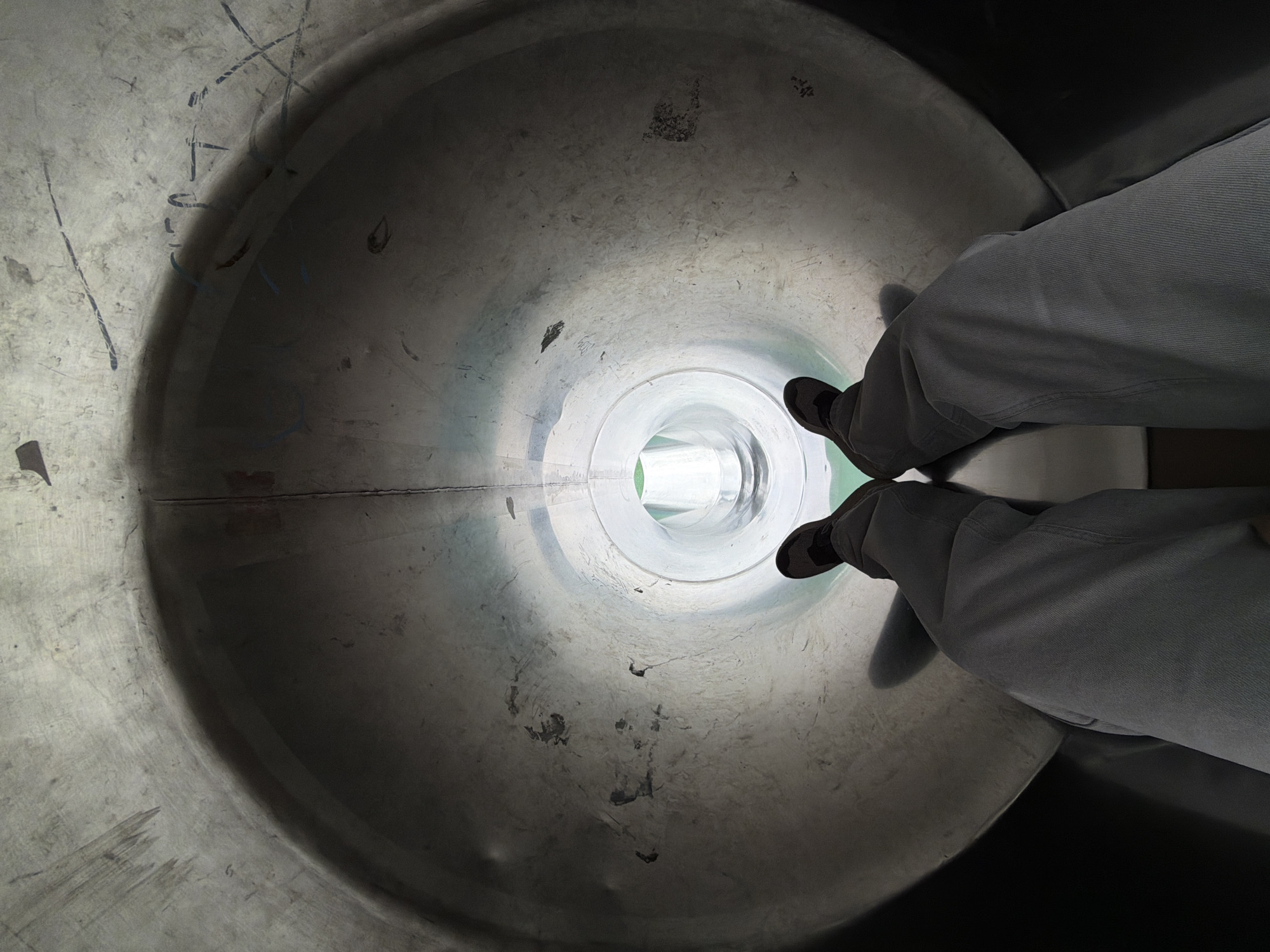 The image shows a person's legs and feet inside a large, silver, tubular slide. The slide appears to be made of metal and has some scratches and marks on its surface. The person is wearing light-colored pants and dark shoes. The perspective is looking down the slide towards a bright opening, suggesting the end of the slide. There is no visible text in the image. The scene is likely an amusement park or playground, given the presence of a slide. The lighting is dim inside the tube, but there is a bright light at the end, indicating daylight outside. The overall mood is neutral, capturing a moment of anticipation or movement.