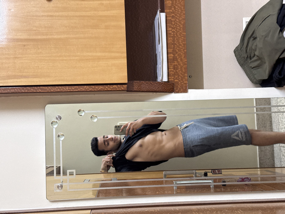 A mirrored selfie captures a man wearing a black t-shirt and gray athletic shorts. His torso is partially exposed. The mirror features decorative elements, including round embellishments and horizontal lines. Above the mirror is a wooden cabinet. To the right, a green and black garment hangs against a light-colored wall. The man is holding a phone in one hand to capture the photo.