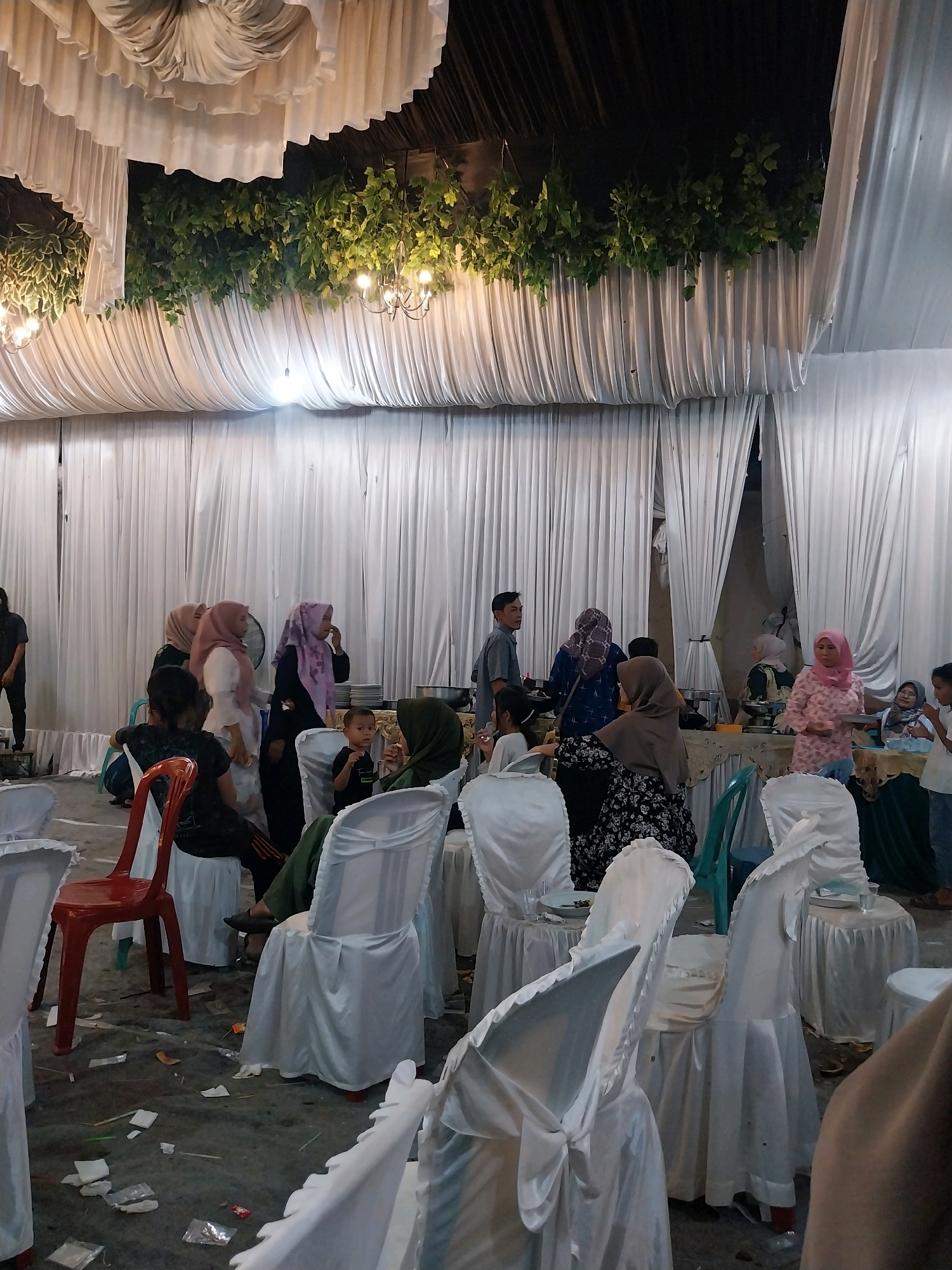 The image depicts an indoor gathering, likely a reception or event, set up under a draped ceiling with white fabric and greenery. The environment appears to be a banquet hall or a similar venue.
In the foreground, several chairs are covered in white fabric, some with bows. The floor is concrete and shows signs of litter, including paper wrappers and other debris. A red plastic chair stands out among the covered chairs, positioned prominently.
A group of people, mostly women wearing hijabs, are gathered around tables in the background. They appear to be interacting, with some holding plates and conversing. There are food service areas with chafing dishes visible on the tables. A young child is seated on one of the covered chairs.
The lighting suggests it is evening or a dimly lit indoor space, with hanging chandeliers contributing to the ambiance. The overall impression is of a social event in progress, with guests mingling and being served.
While the image itself does not provide explicit location markers, the provided context indicates the scene is located in the city of Cirebon, Indonesia.
