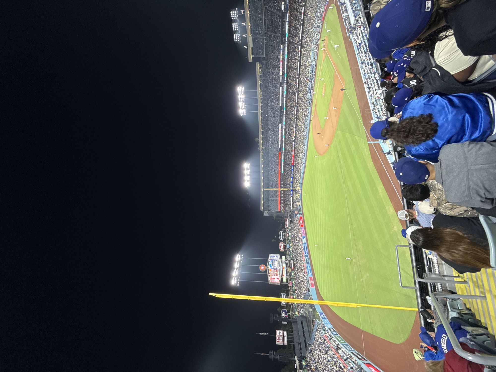 The image shows a baseball game at night from an elevated perspective. The field is illuminated by stadium lights, and the stands are full of spectators. Many people are wearing blue clothing and baseball caps, indicative of support for the Los Angeles Dodgers. In the foreground, portions of the crowd are visible, including individuals in Dodger apparel. The stadium's outfield wall and advertising boards are visible along the field's perimeter. The scene is set in Los Angeles, United States of America, based on the team colors and stadium location.