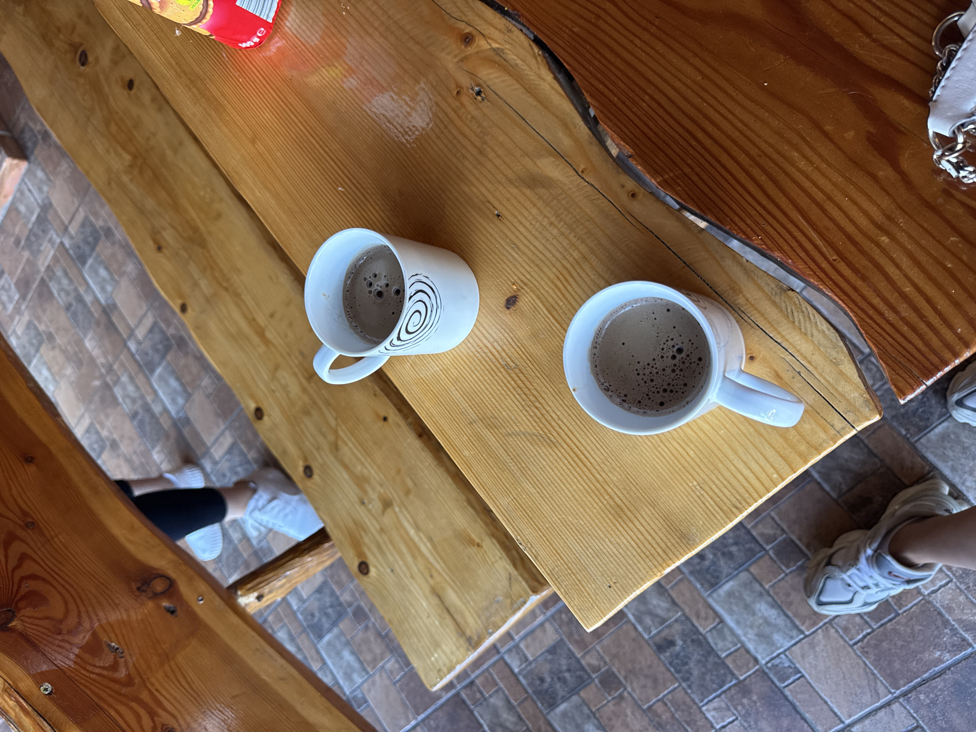 The image depicts two white ceramic mugs filled with a frothy brown beverage, likely coffee or hot chocolate, placed on a rustic wooden table. The mugs are positioned on the table's surface, which is made of several wooden planks, exhibiting a natural, unfinished grain. One mug features a black spiral pattern on its side. In the background, through a gap in the wooden seating, a person's legs wearing black pants and white sneakers are visible, standing on a tiled floor. Another person's feet, wearing white sneakers, are visible in the lower left corner of the image, on the same tiled flooring. To the right of the mugs, a portion of a red and yellow package, possibly containing biscuits, is visible on the table. The setting appears to be an outdoor or semi-outdoor area, with natural light illuminating the scene. The location is identified as Cârțișoara, Romania.