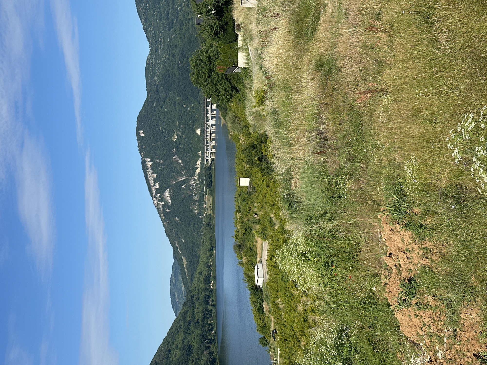 A wide, outdoor shot captures a serene landscape featuring a calm body of water surrounded by verdant, rolling hills under a clear blue sky. In the foreground, tall, dry grasses and a patch of reddish-brown soil dominate the scene, interspersed with small white wildflowers.
To the left, the water stretches across the frame, bordered by a dense line of green bushes and trees. A small, white rectangular building with a dark roof sits near the edge of the water, accompanied by a single black car parked on a paved area.
Further back, on the opposite side of the water, a long, white bridge with numerous supports spans across the expanse. The hills in the background are covered in lush green foliage, with some areas revealing rocky cliff faces. The sky above is a bright blue with a few wispy white clouds scattered across it.
The scene appears to be a natural landscape with some man-made structures, possibly a reservoir or lake with a bridge and a small building. There are no people visible in the image. The overall impression is one of tranquility and natural beauty, with the elements of water, land, and sky harmoniously coexisting.