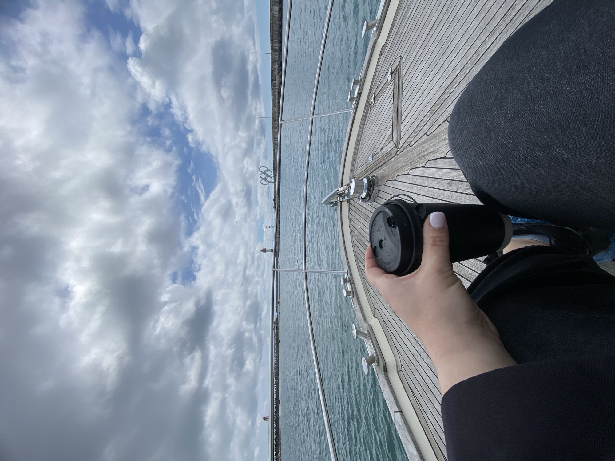 Here is a detailed description of the image:
**Scene Overview:**
The image appears to be a first-person perspective taken on the deck of a boat or yacht. The view is oriented towards the horizon, where the sea meets the sky, with some cityscape visible along the coast. The weather seems overcast with a cloudy sky dominating the upper portion of the picture.
**Location:**
The scene is identified as Sochi, Russia. The presence of the Olympic rings monument on the coast in the distance is a distinctive landmark associated with the city of Sochi, which hosted the 2014 Winter Olympics.
**Objects:**
* **Boat/Yacht:** The image is taken from the deck of a boat, evident from the wooden planking of the deck and the presence of nautical hardware like railing supports.
* **Coffee Cup:** A black travel coffee cup is held in the foreground by a person's hand.
* **Clothing:** The person is wearing black clothing, likely a jacket or sweater, and possibly dark pants.
* **Sky:** Overcast with large clouds.
* **Water:** Calm waters of the Black Sea.
* **Cityscape:** Elements of the city of Sochi, including buildings and possibly infrastructure, are visible along the distant coastline.
* **Olympic Rings:** The Olympic rings monument stands tall on the coast.
**Subjects & Actions:**
* **Person Holding Cup:** The only human element is the arm and hand of a person holding a black coffee cup. The hand has light skin, and the fingernails are painted with white nail polish.
* **Sipping/Holding:** The action is of someone either about to drink from or holding the coffee cup.
**Environment:**
The environment suggests a leisure activity on the sea. The day appears to be cloudy but otherwise calm. The location is near the coast of a city with distinctive landmarks.
