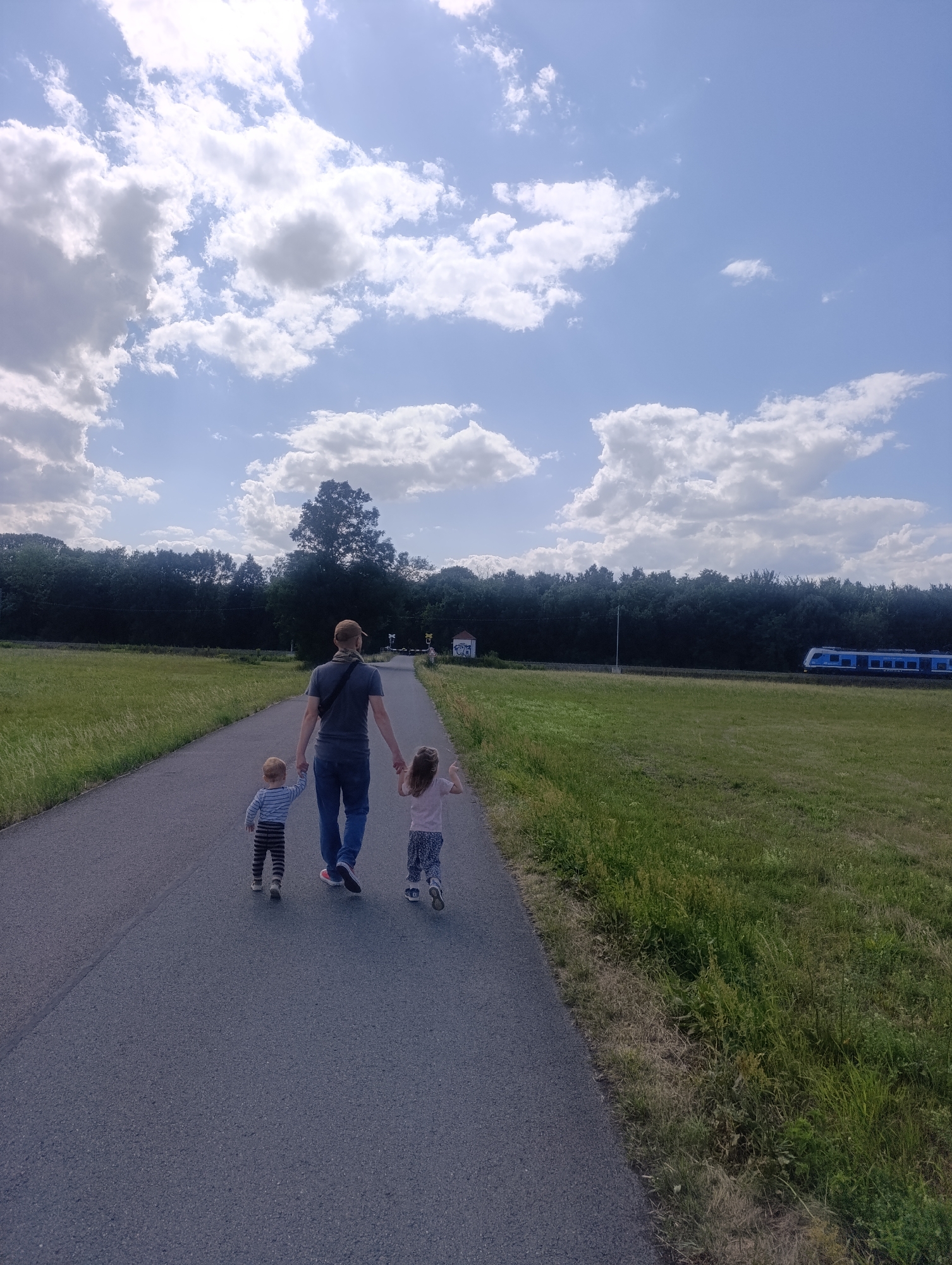 In Olomouc, Czechia, a father walks hand-in-hand with his two young children on a paved path. The father, wearing a gray t-shirt, blue jeans, and a baseball cap, holds hands with his son on the left, who is dressed in a striped long-sleeved shirt and striped pants, and his daughter on the right, wearing a pink t-shirt and patterned pants. They are walking away from the camera on a path bordered by grassy fields. In the background, a dense line of trees runs horizontally across the image, with a blue and white train visible on tracks to the right of the trees. The sky above is bright blue with scattered white clouds. The overall impression is a peaceful, outdoor scene.